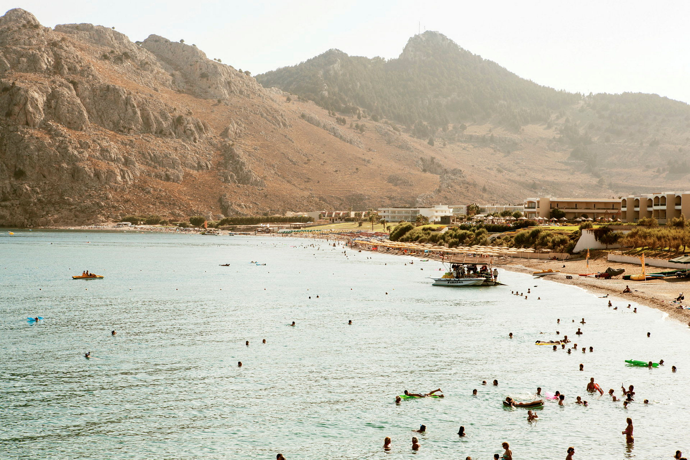 En strand ved en bugt omgivet af bjerge, med personer der svømmer i vandet og soler sig på stranden. (AI-genereret tekst)