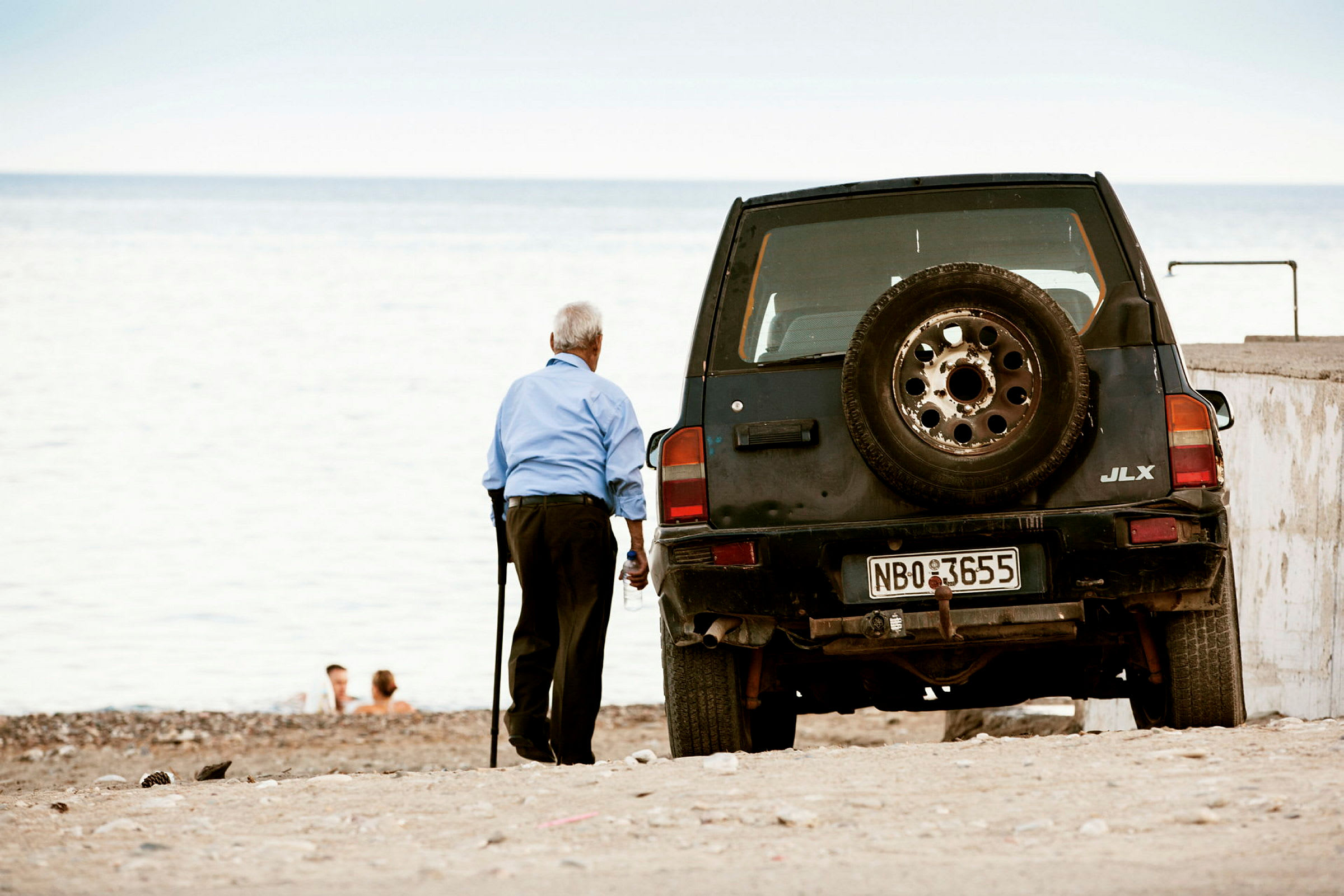 En ældre person står på en strand ved siden af en sort bil med reservehjul bagpå. (AI-genereret tekst)