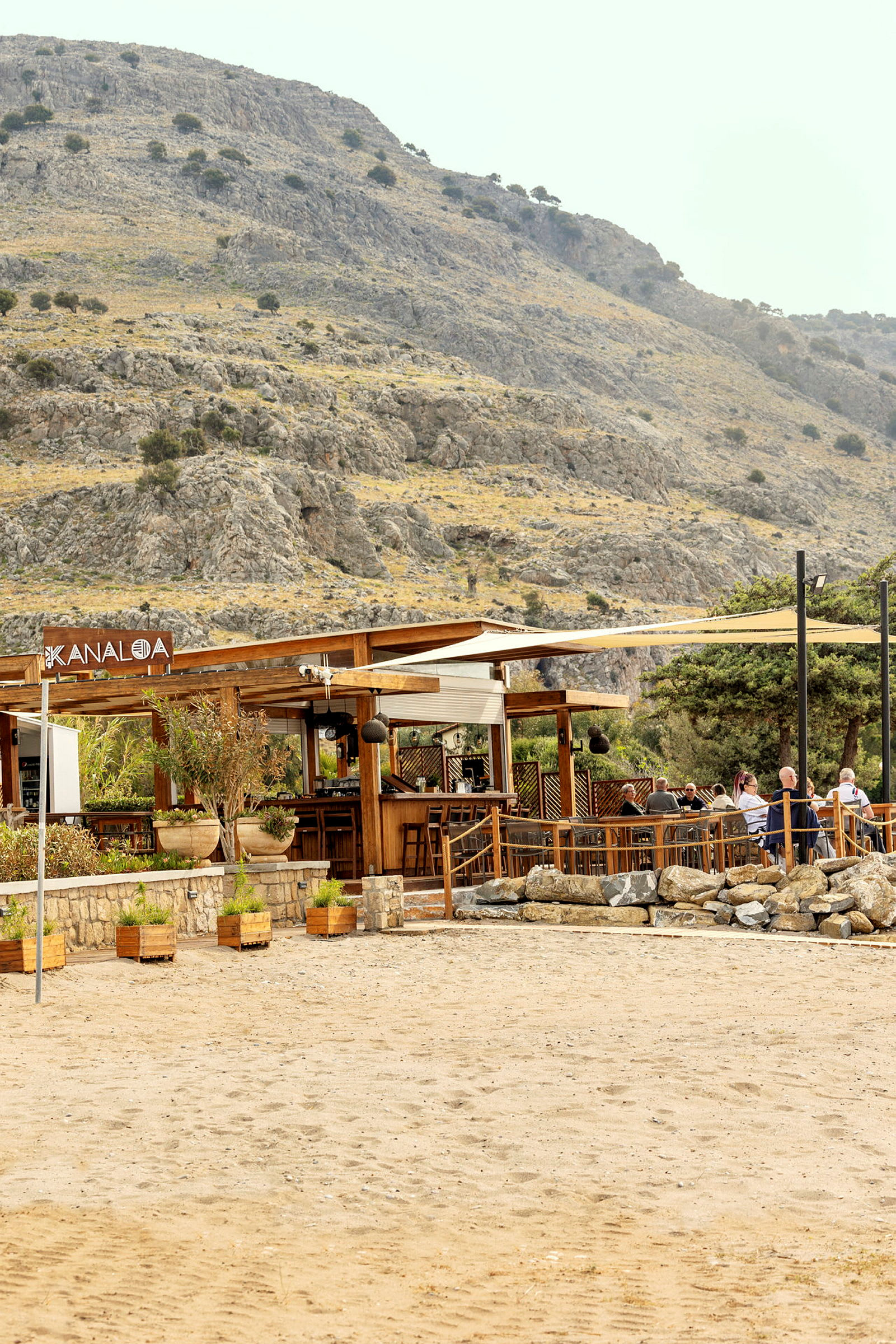 En strandbar med træmøbler og parasoller, omgivet af en bjergrig kulisse. (AI-genereret tekst)