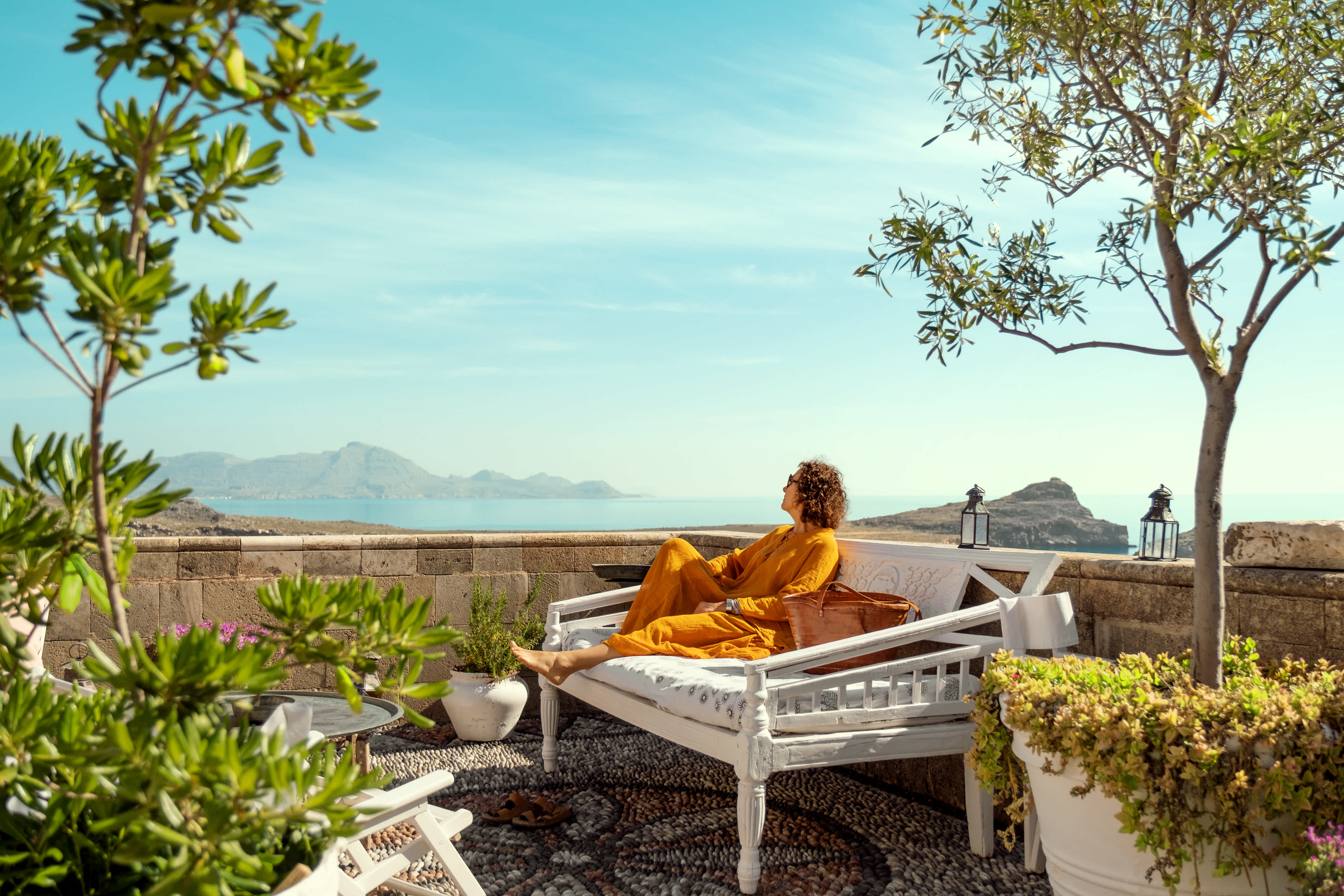 En person slapper af på en hvid bænk på en terrasse omgivet af grønt med udsigt over havet og bjergene. (AI-genereret tekst)