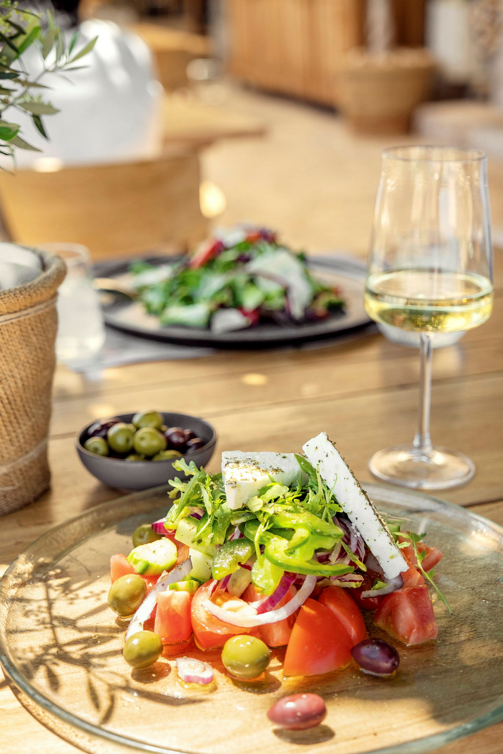 Billede af en tallerken med salat, fetaost og oliven samt et glas hvidvin på et træbord. (AI-genereret tekst)
