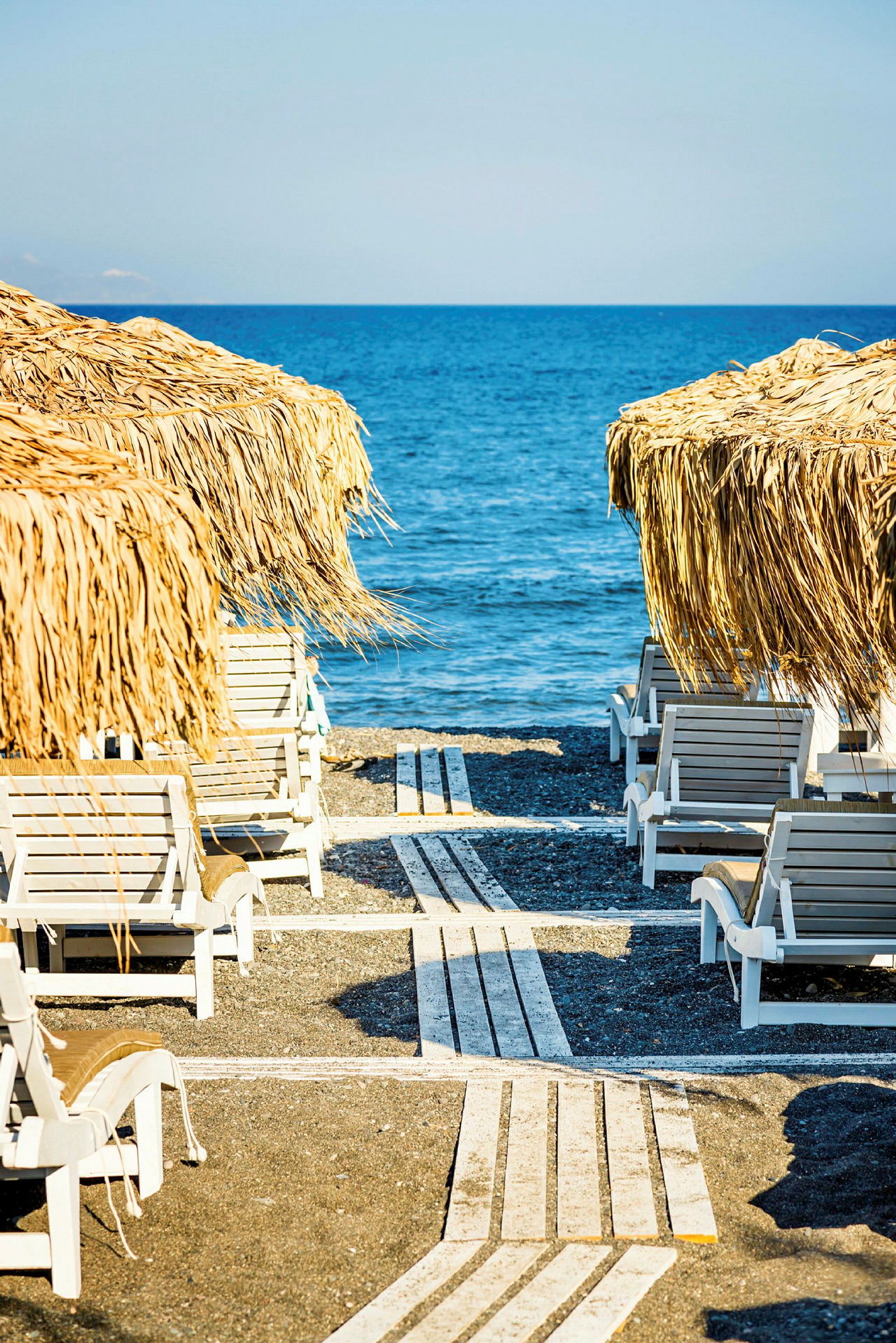 Solstole på en strand med parasoller af plantemateriale og havet i baggrunden. (AI-genereret tekst)