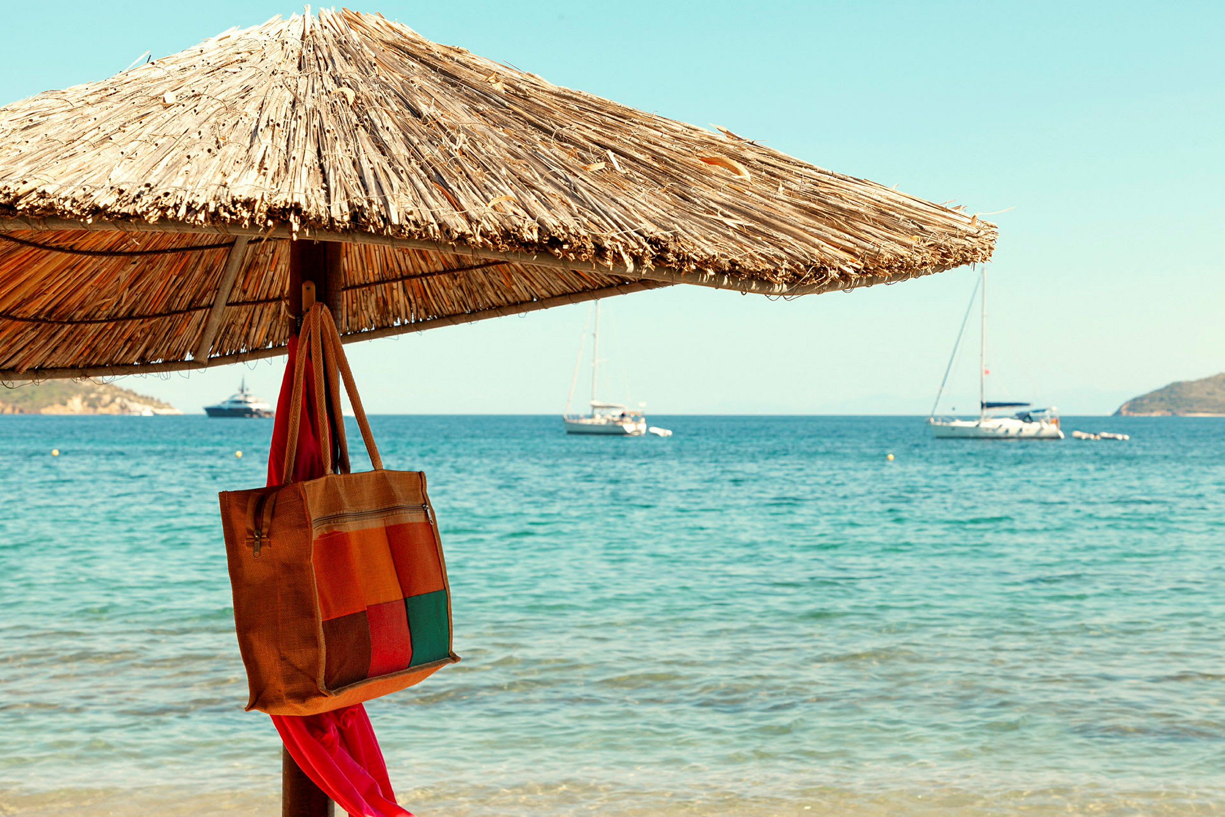 Billede af en strand med en parasol lavet af halm. En taske hænger på parasollen, og sejlbåde er tydelige ude på havet. (AI-genereret tekst)