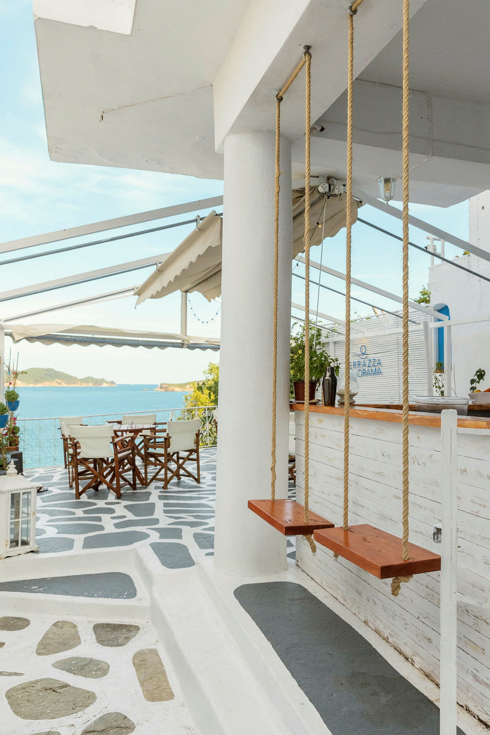 Billede af en terrasse med stole og borde, havudsigt og to trægynger foran en søjle. (AI-genereret tekst)