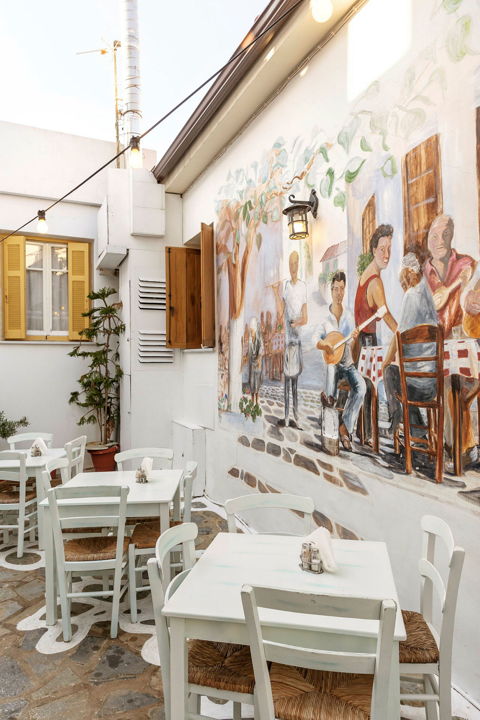 Udendørsområde med hvide borde og stole, væg dekoreret med maleri af mennesker i en restaurantmiljø. (AI-genereret tekst)