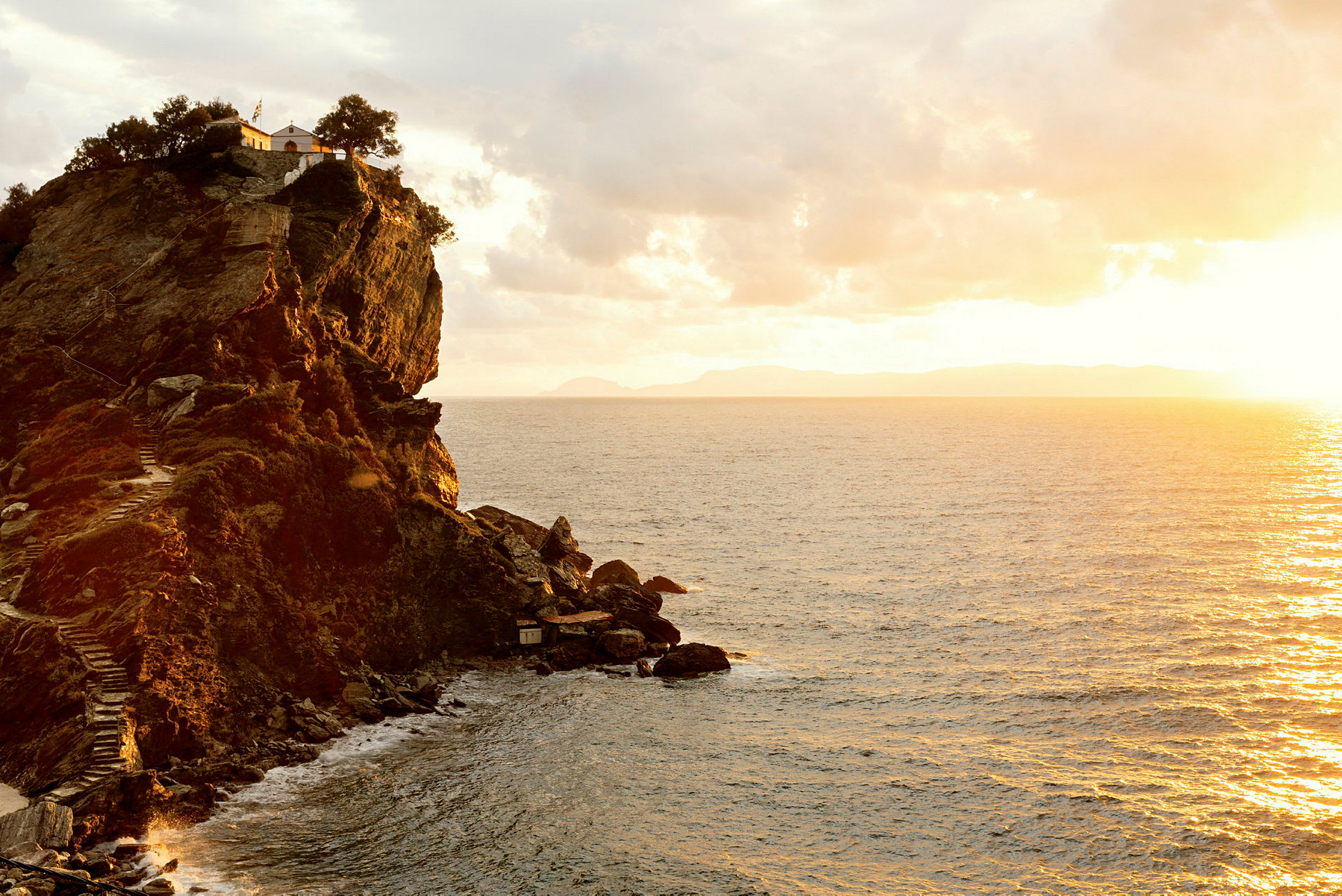 En klippe ved kysten med en bygning og træer på toppen. Havet ses under en solnedgang. (AI-genereret tekst)