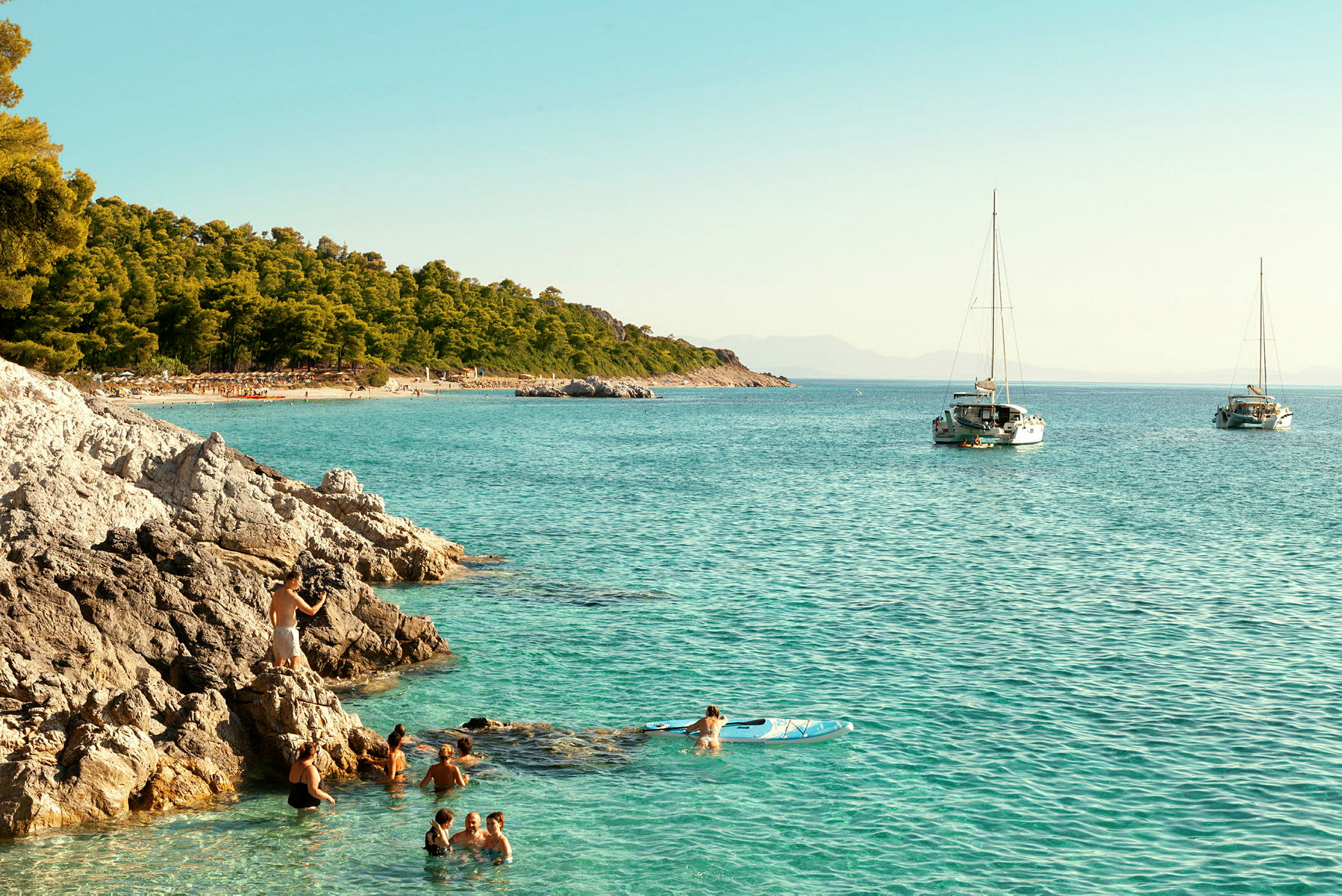 Billedet viser en strand med klipper og klart blåt vand. Folk bader, og en person flyder på et paddleboard. To sejlbåde er fortøjret længere ude på vandet. (AI-genereret tekst)
