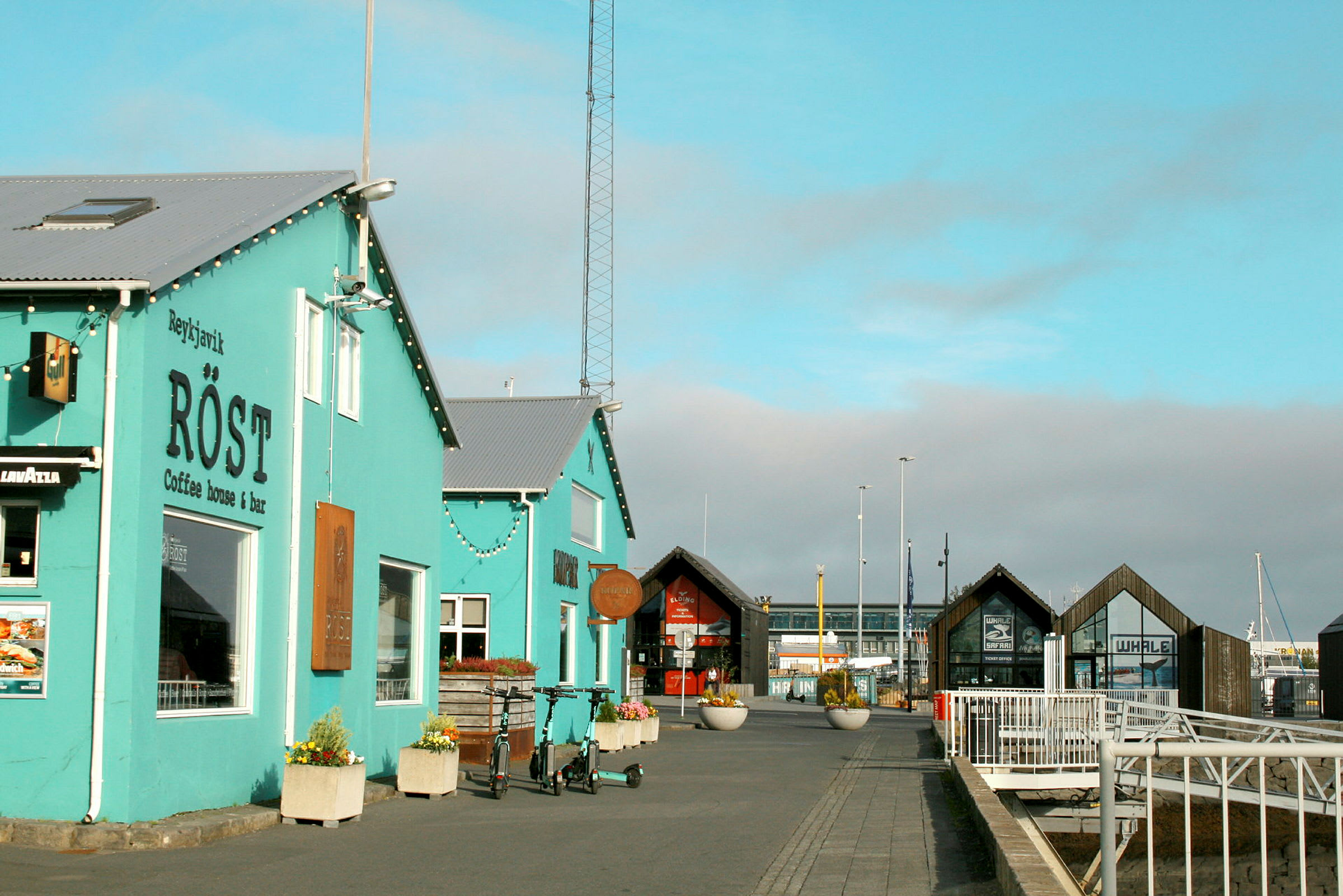 Et turkis caféhus ved en havn med bygninger og trædæk i baggrunden. (AI-genereret tekst)