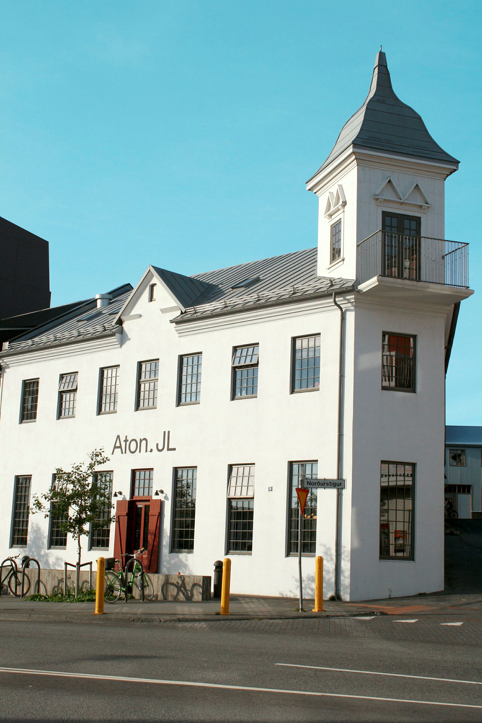 En hvid bygning med et tårn og store vinduer. Bygningen har et skilt der siger "Aton.JL", og der er cykler parkeret foran. (AI-genereret tekst)