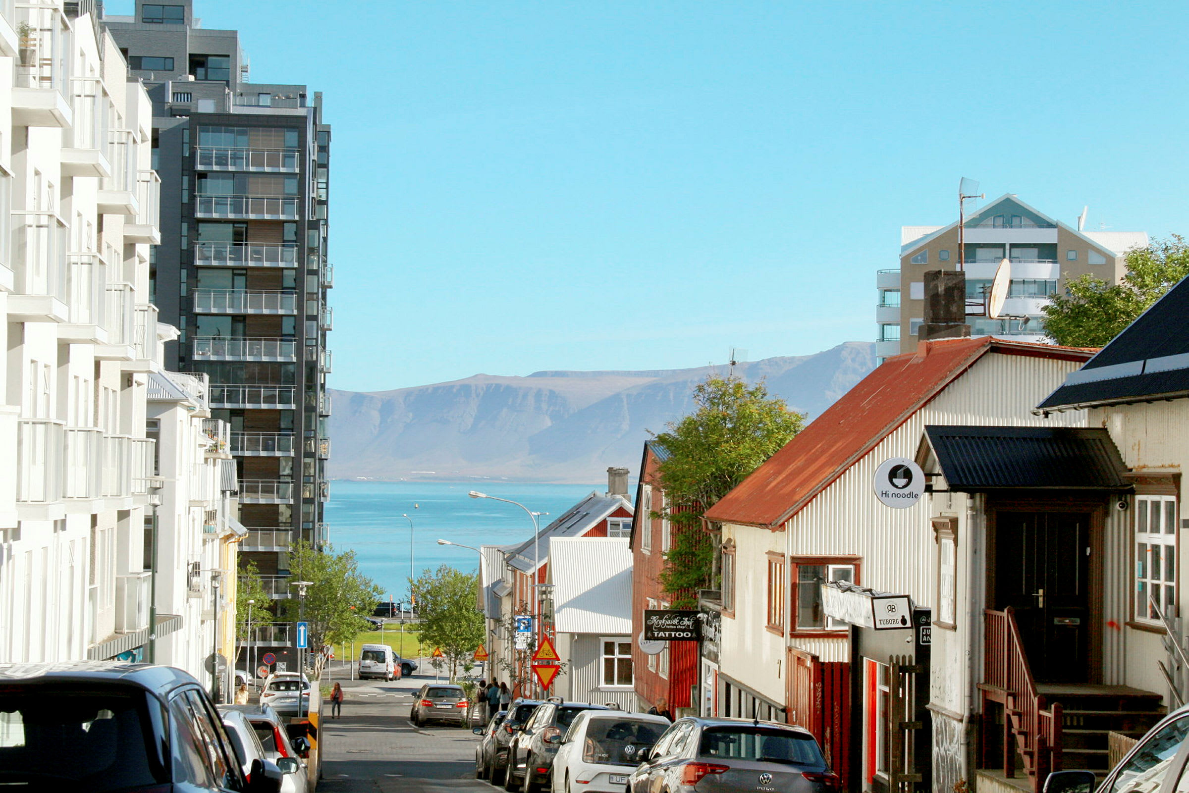 En gade i en by med bygninger på begge sider og udsigt over havet og bjerge i baggrunden. (AI-genereret tekst)