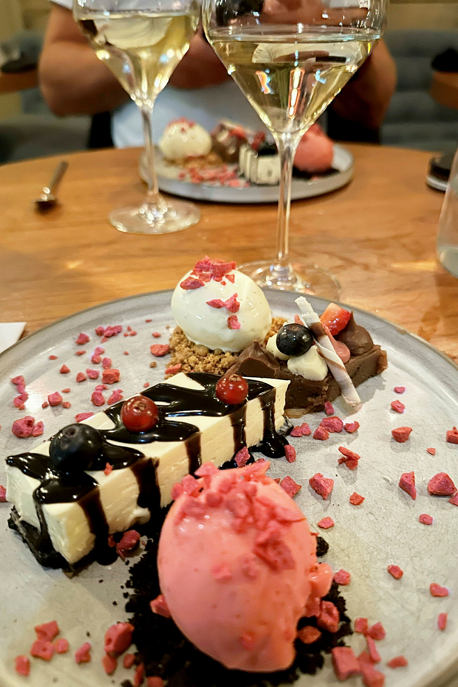 En tallerken med forskellige desserter, herunder en lyserød sorbet, ostekage og andre små søde sager, pyntet med bær og sauce. To glas hvidvin kan ses i baggrunden. (AI-genereret tekst)