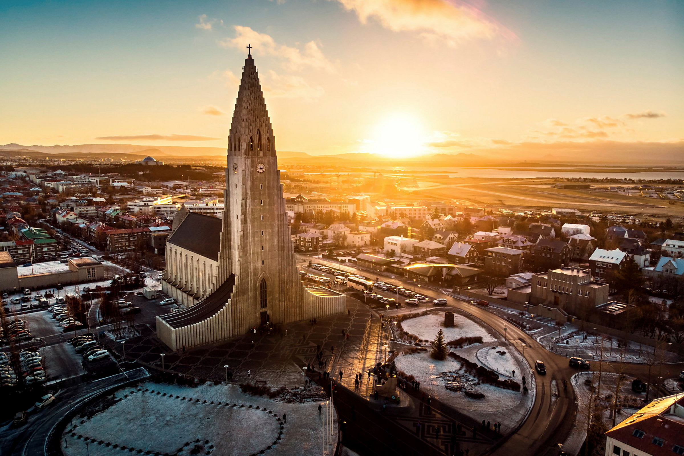 En kirke med moderne arkitektur og en høj tårnstruktur, omgivet af en by og et landskab under en solnedgang. (AI-genereret tekst)