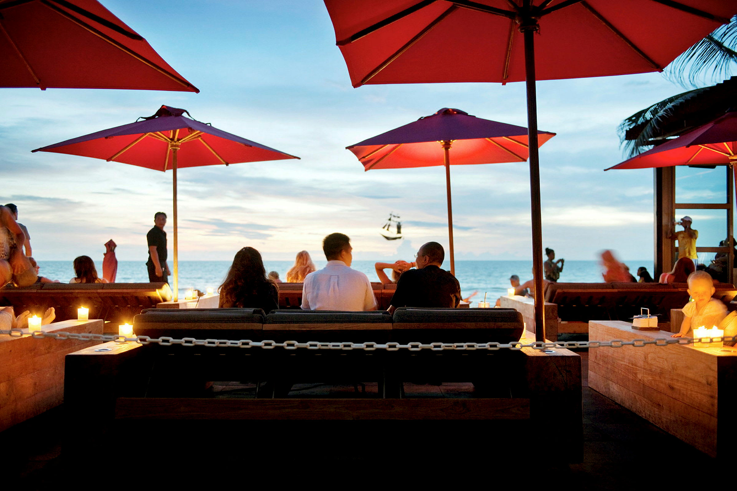 Folk sidder ved et bord på en udendørs restaurant med røde parasoller og havet i baggrunden. (AI-genereret tekst)
