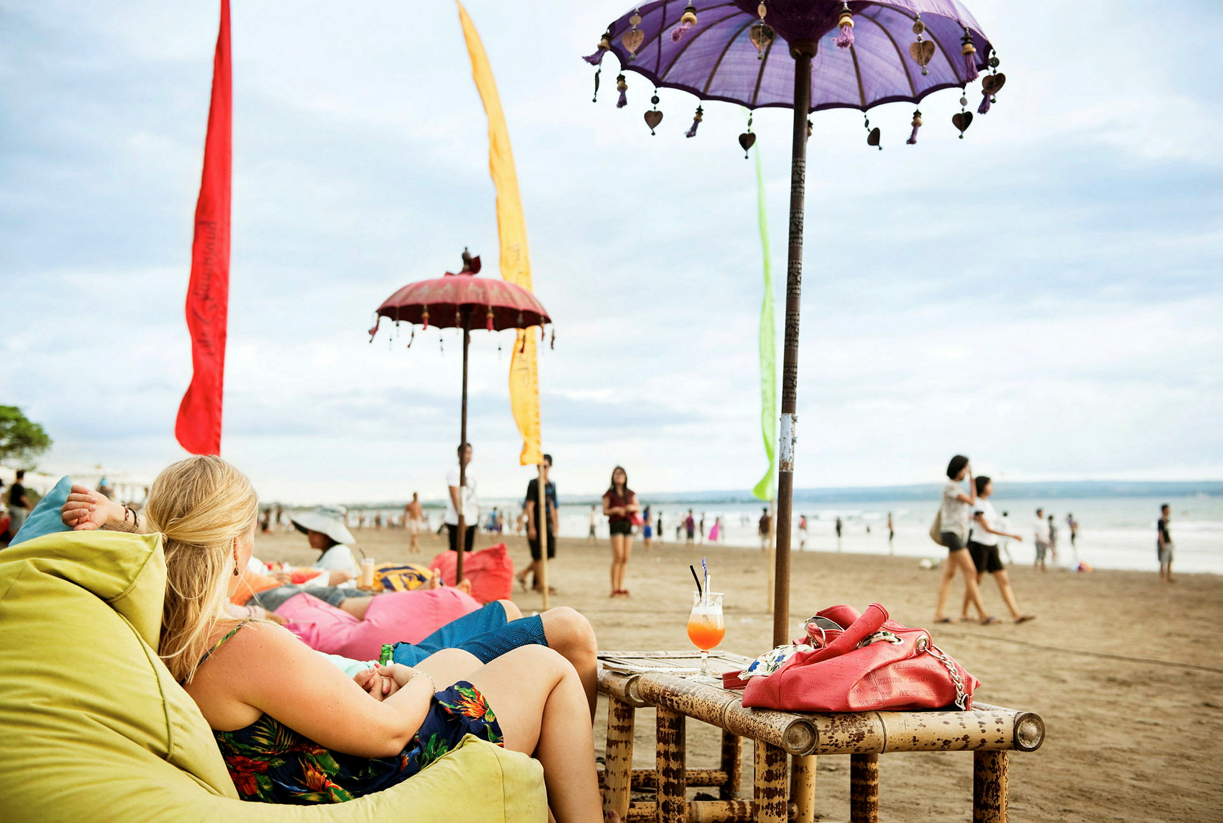 Strand med personer, der sidder i farverige sækkestole under dekorerede parasoller. I forgrunden står et bambusbord med en drink og en rød taske. (AI-genereret tekst)