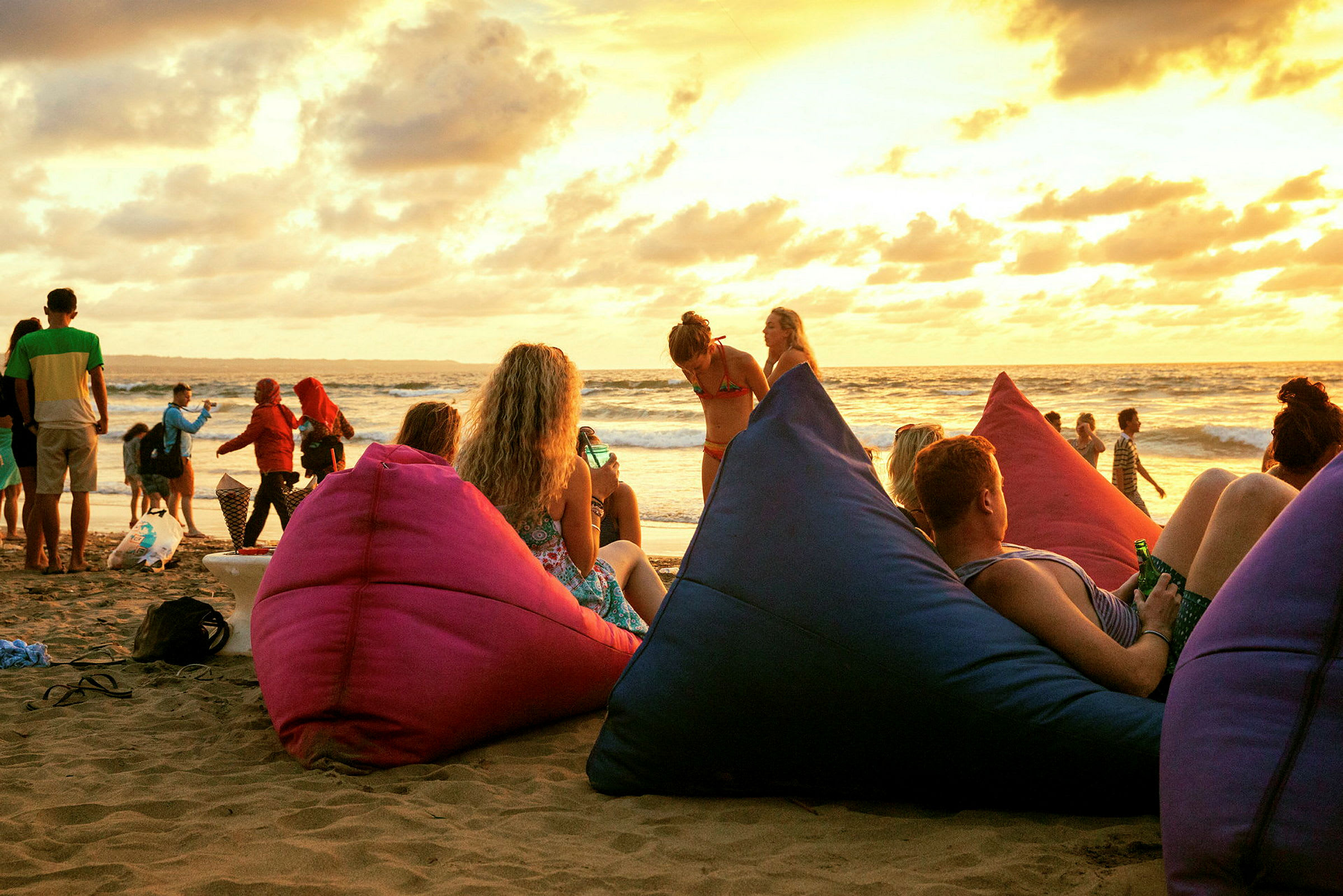Afslappet strandscene ved solnedgang med personer der sidder på farverige sækkestole. (AI-genereret tekst)