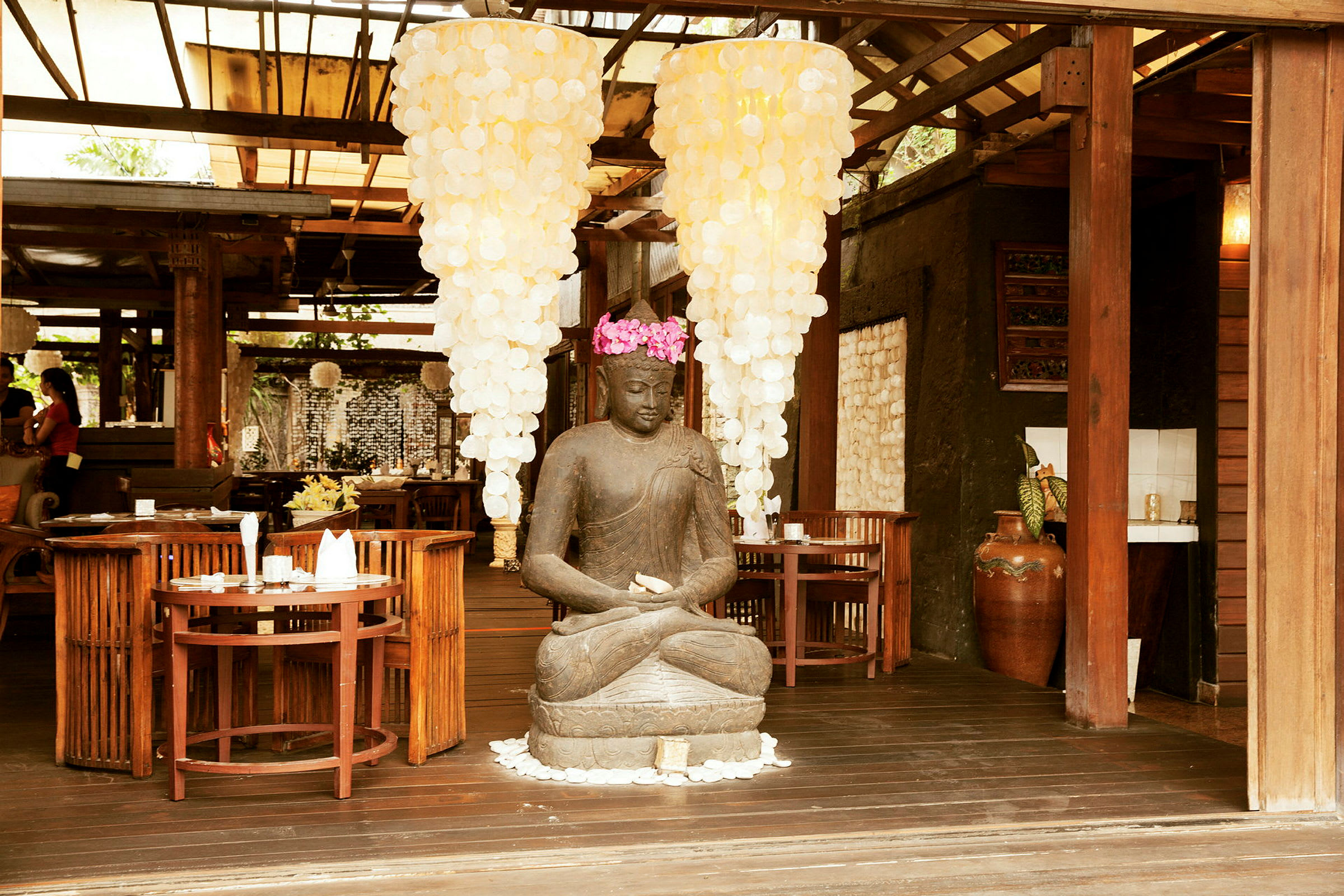 Statue af siddende Buddha omgivet af træborde og stole, med store lysekroner hængende ovenover. (AI-genereret tekst)