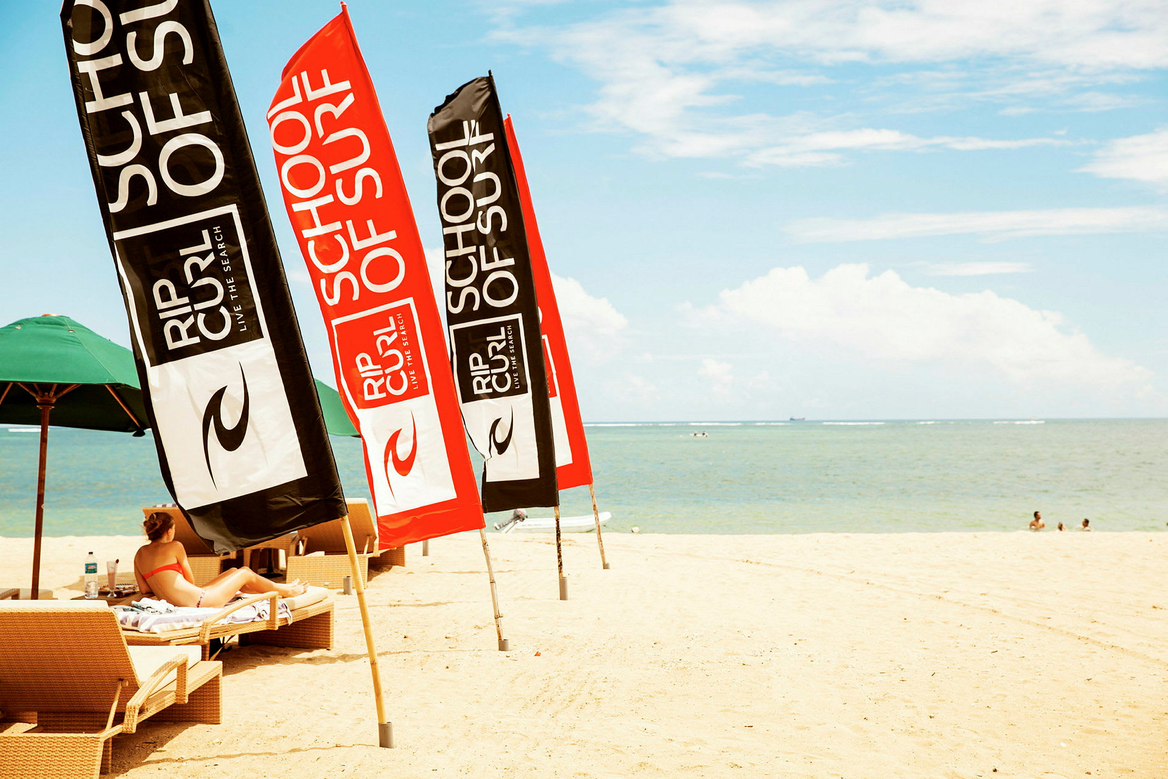 Strand med liggestole og flag for surfskoletilbud nær havet. (AI-genereret tekst)