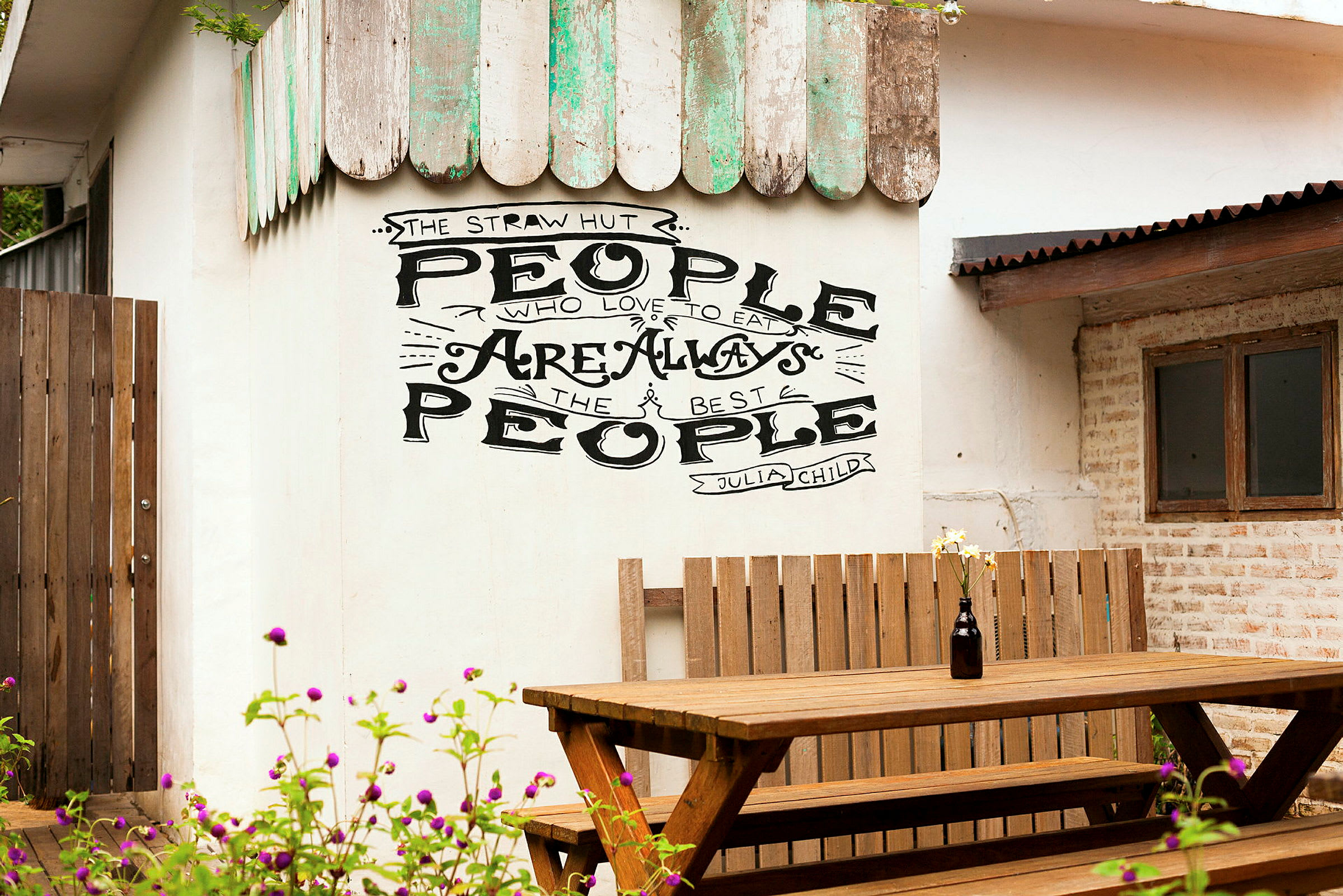 Væg med teksten 'People who love to eat are always the best people' over en træbænk og et bord. (AI-genereret tekst)