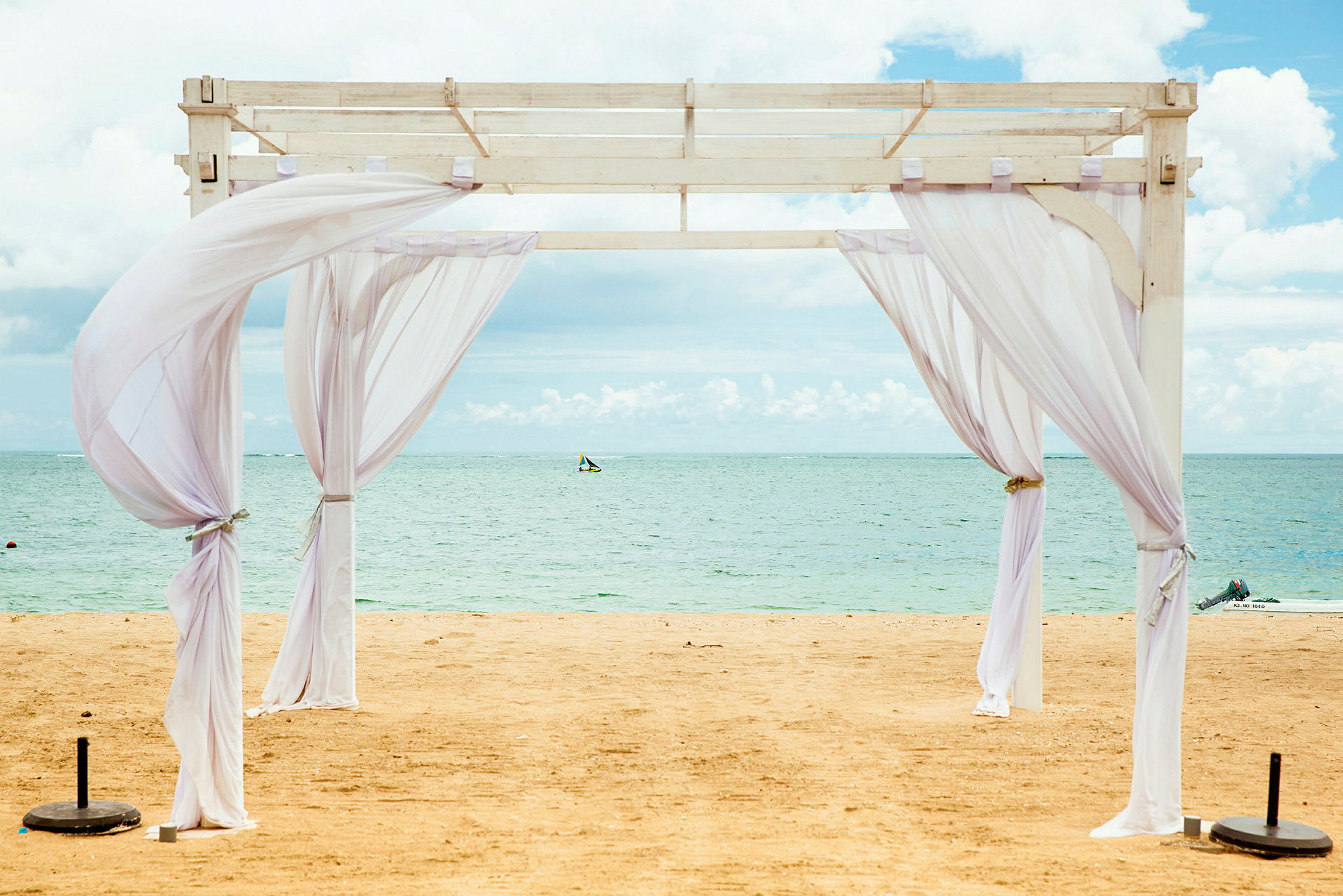 Billedet viser en hvid pergola med tynde stoffer på en sandstrand ved havet. (AI-genereret tekst)