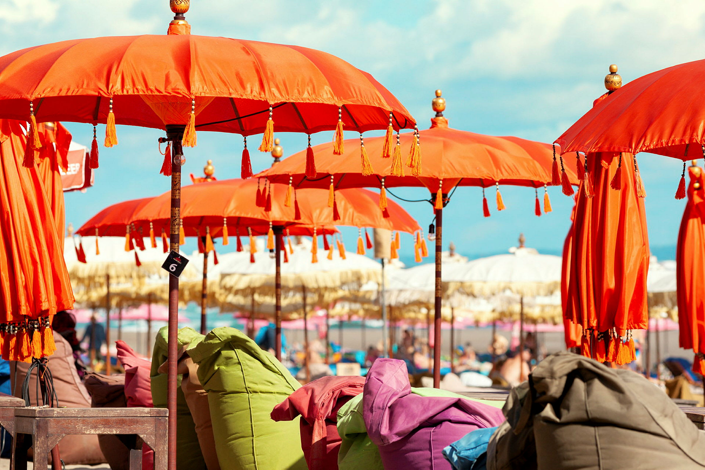 Flere orange parasoller på række med farverige siddehynder under imod en strandscenes baggrund. (AI-genereret tekst)