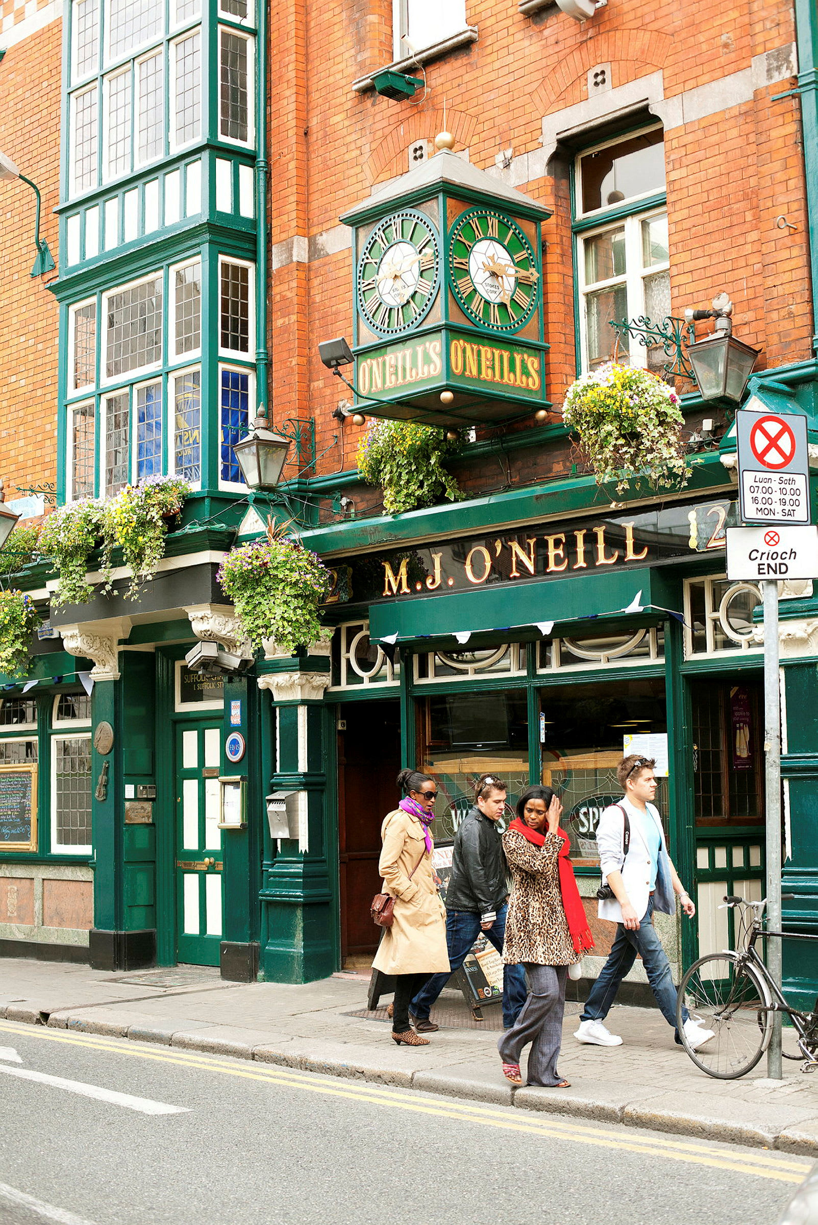 En traditionel irsk pub med murstensvægge og et grønt klokketårn over indgangen. Flere personer går forbi udenfor. (AI-genereret tekst)