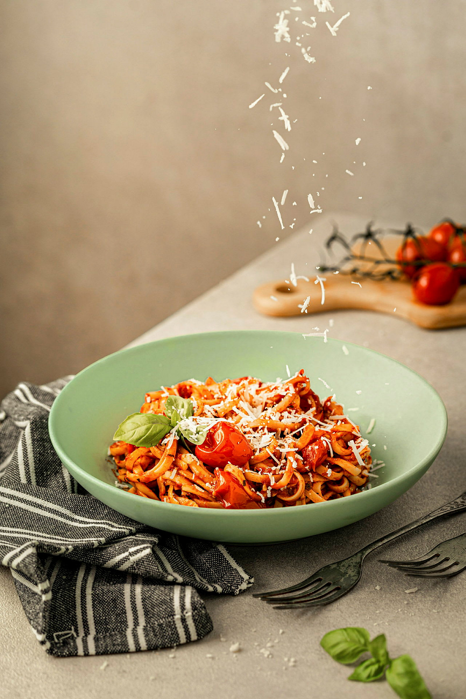 En tallerken med pasta, toppet med tomater, basilikum og revet ost, placeret på en grå overflade med bestik og en sort-hvid klud. (AI-genereret tekst)