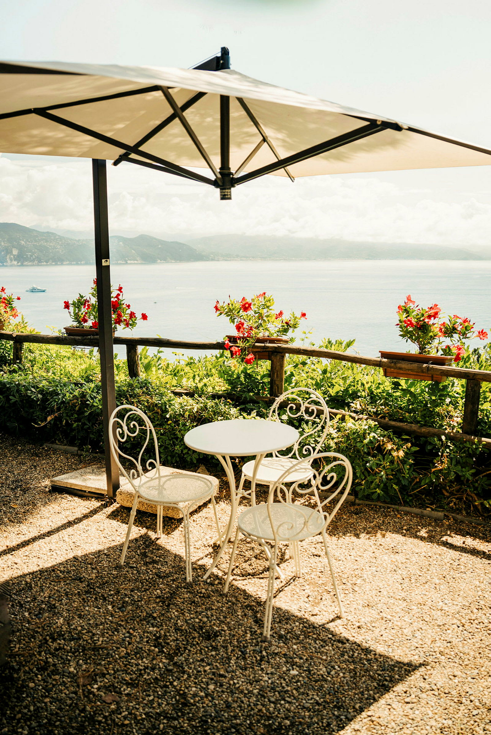 Et bord og to stole placeret under en parasol med udsigt over havet, omgivet af blomstrende planter. (AI-genereret tekst)