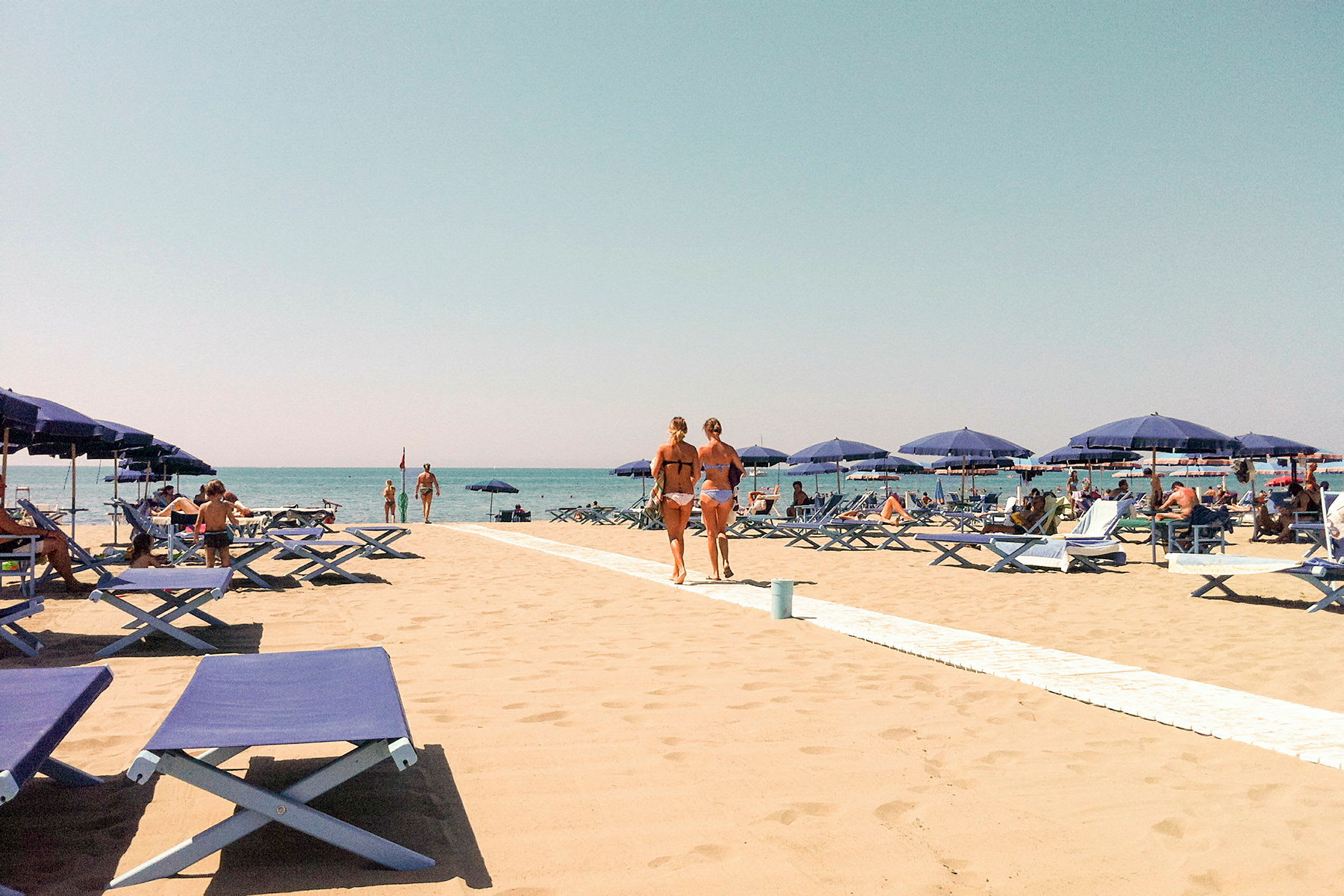 En sandstrand med blå liggestole og parasoller. Folk soler sig og går på stranden med en klar blå himmel i baggrunden. (AI-genereret tekst)