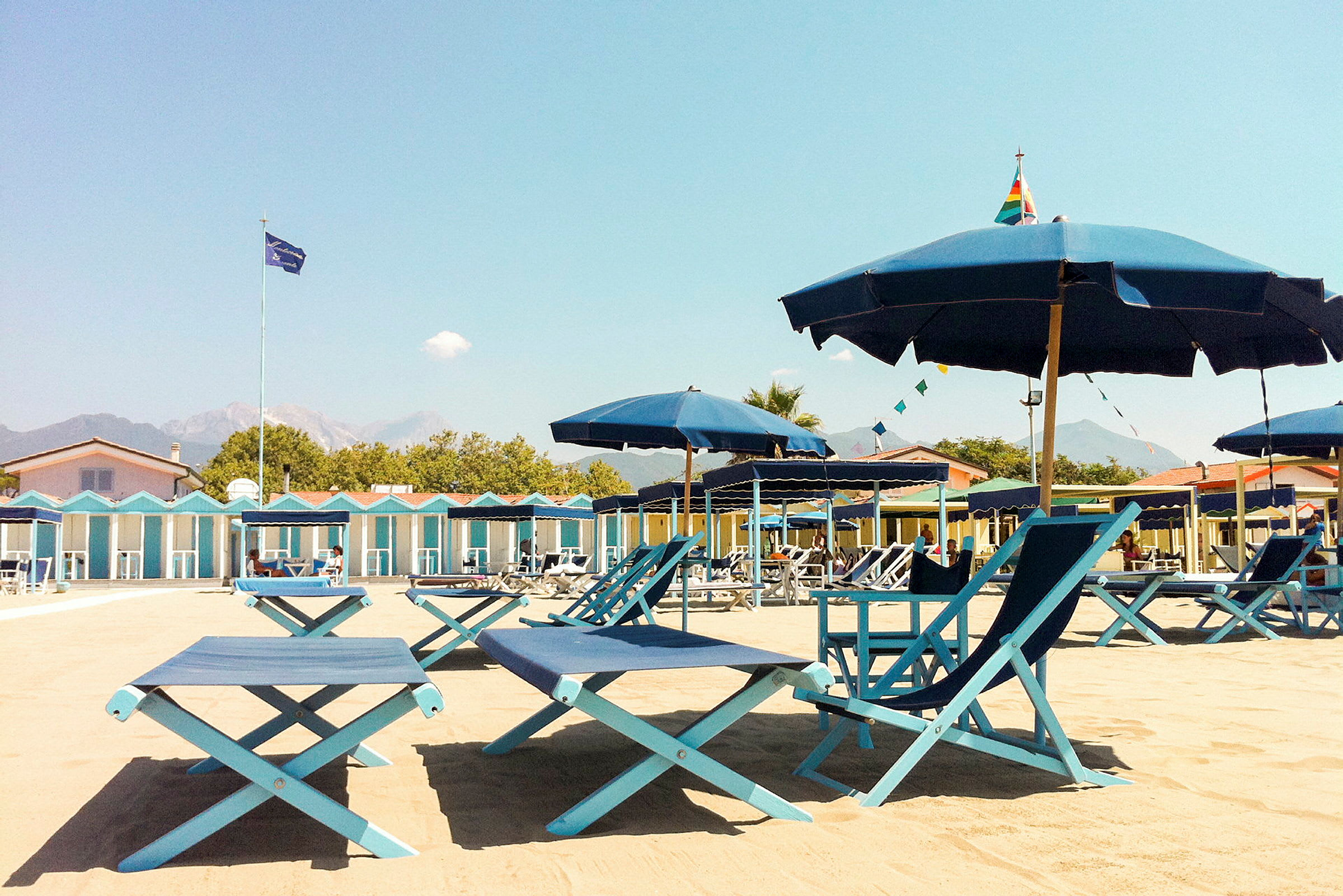 Strandmiljø med liggestole og parasoller samt et område med strandhytter i baggrunden. (AI-genereret tekst)