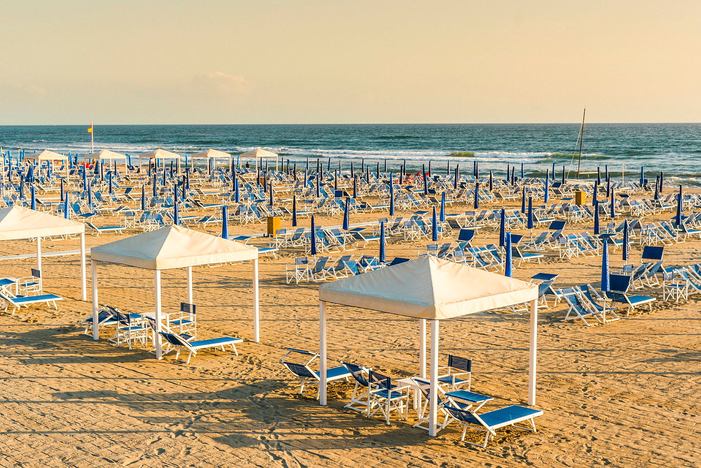 En sandstrand med opstillet blå og hvide liggestole samt parasoller. I forgrunden er der cabanas med siddegrupper. (AI-genereret tekst)