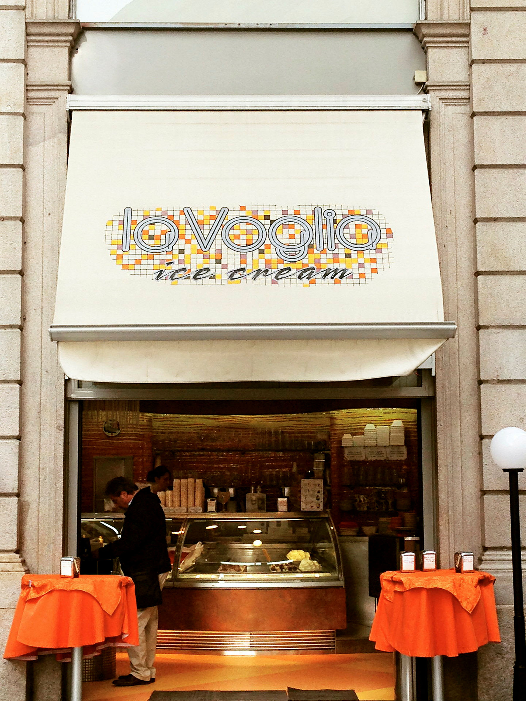 En isbutik med et skilt, der siger "La Voglia ice cream" og to små borde med orange duge foran indgangen. (AI-genereret tekst)