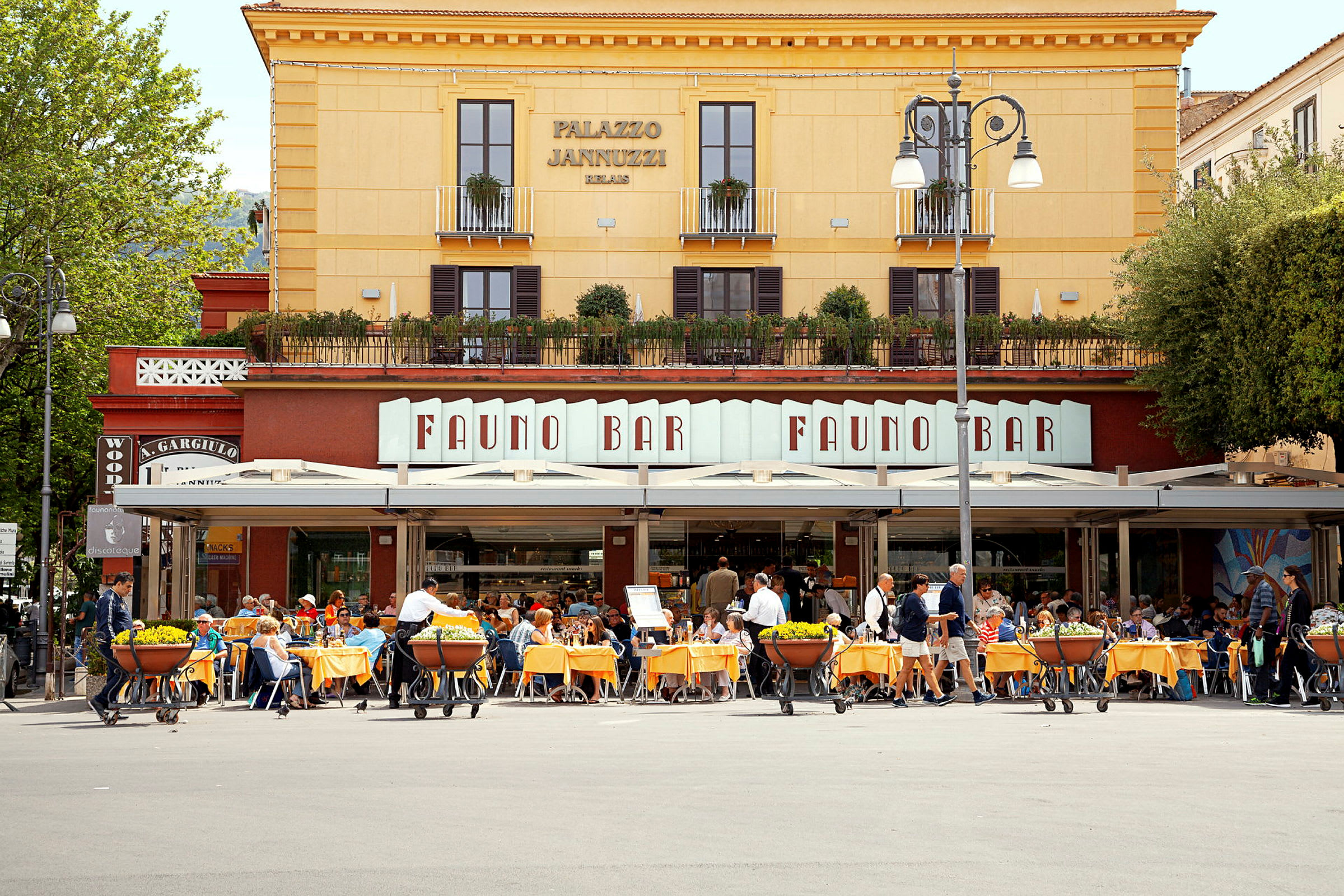 En restaurant med udendørs servering foran en gul bygning med skilte, der viser navnet 'Fauno Bar'. (AI-genereret tekst)