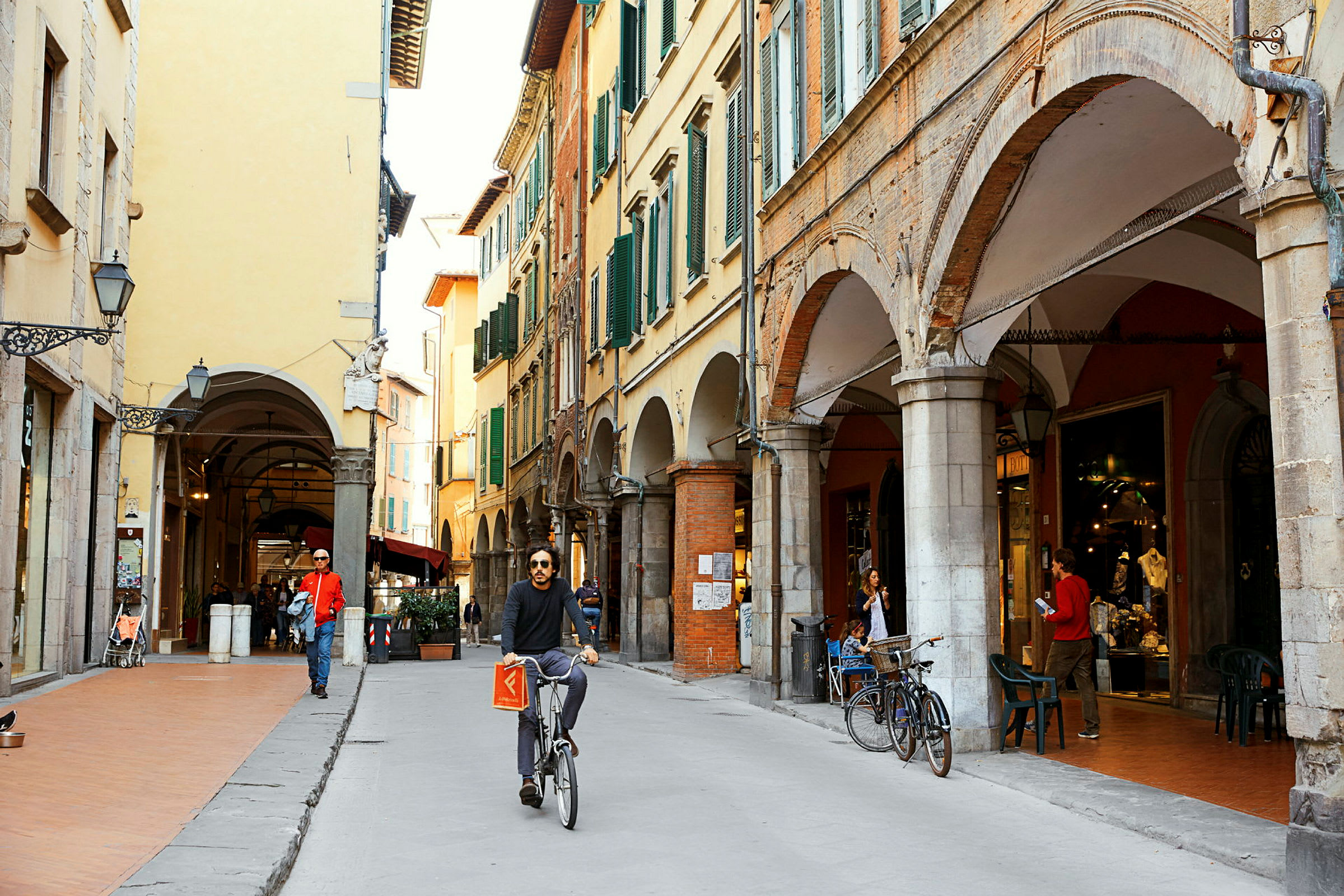 Gade med historiske bygninger og arkader. En person cykler forbi, og nogle personer går forbi eller sidder ved en café. (AI-genereret tekst)