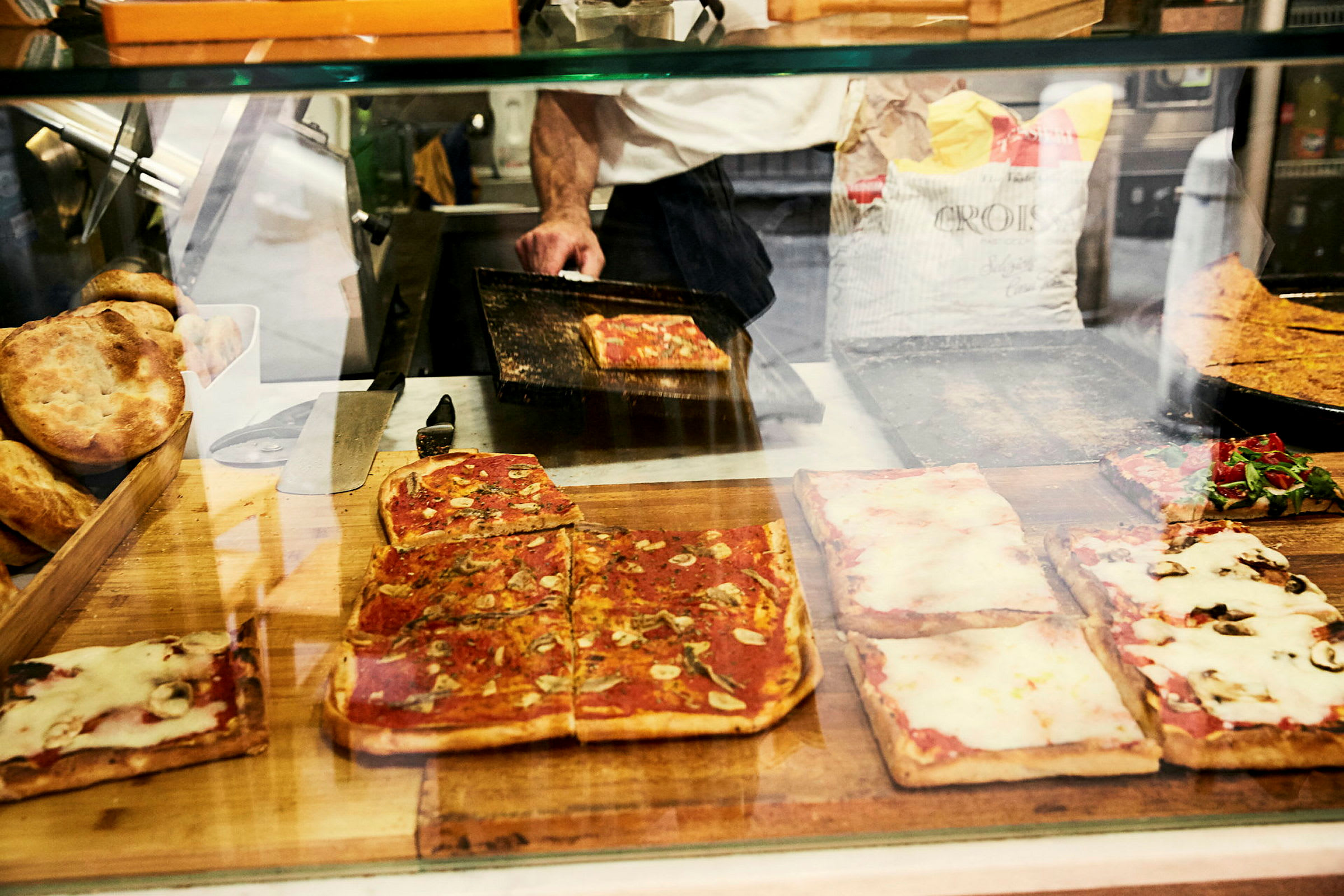 Firkantede pizzaer med forskellige toppings bag en glasskærm, samt brød og en pizzaovn i baggrunden. (AI-genereret tekst)