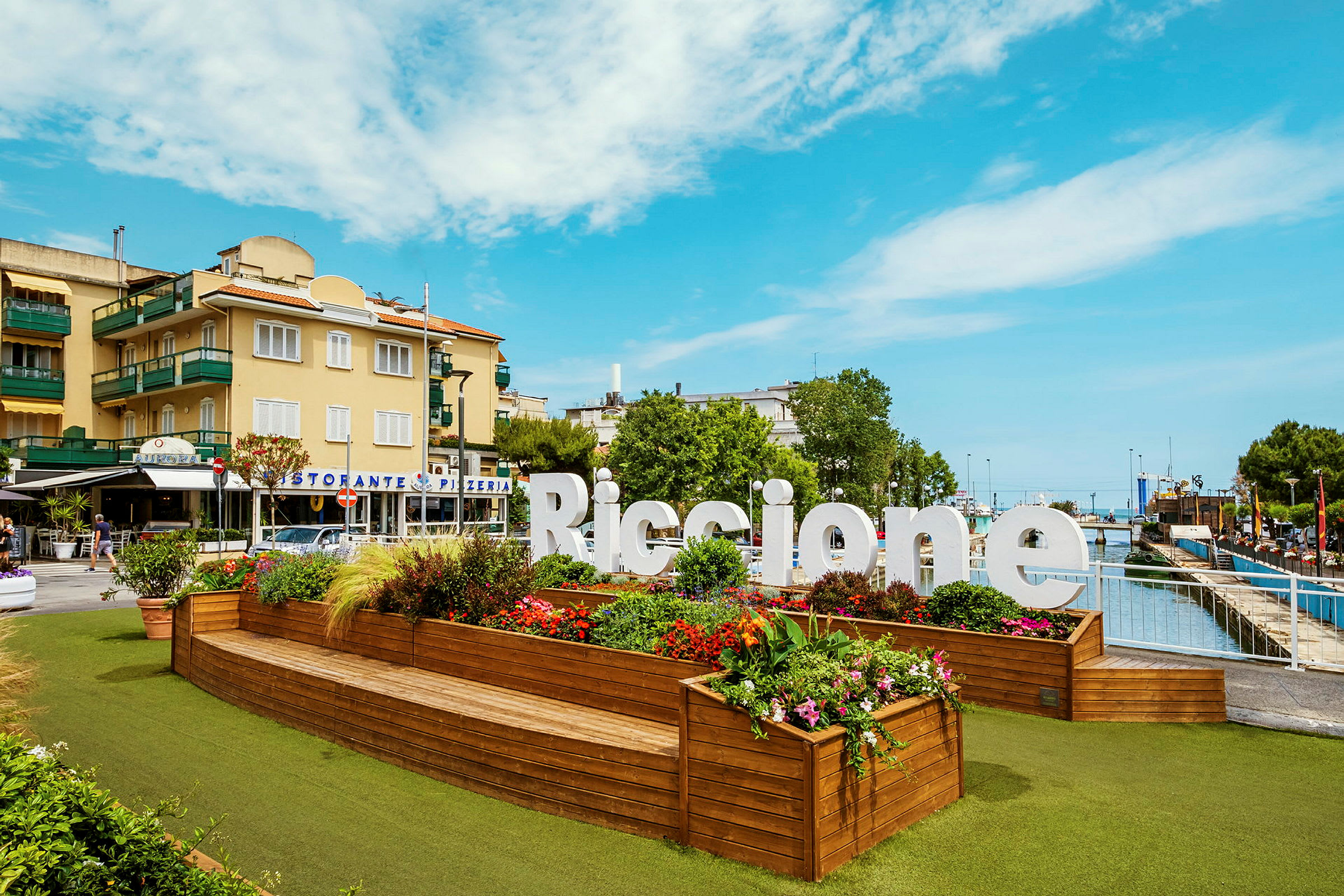 Et stort skilt med teksten "Riccione" står foran et planteområde med farverige blomster og grønne planter. I baggrunden ses bygninger og en klar himmel. (AI-genereret tekst)