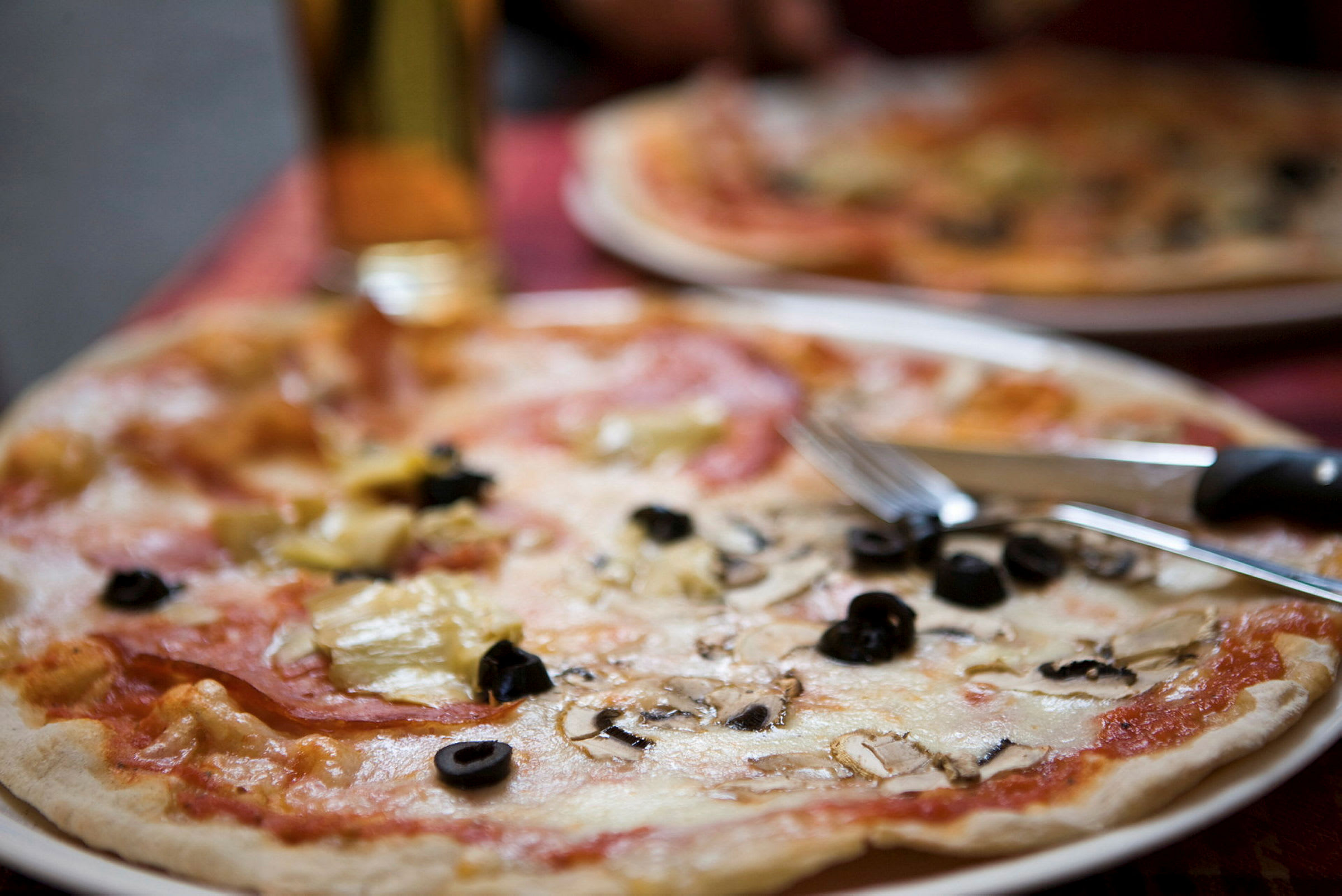 En tallerken med pizza toppet med ost, sorte oliven og champignoner. Bestik ligger på kanten af tallerkenen. (AI-genereret tekst)