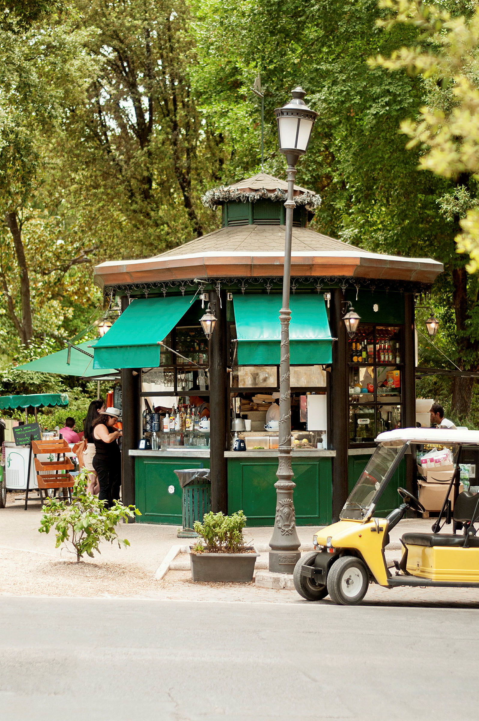 Grøn kiosk med markiser og omkringliggende træer, en gul golfbil er synlig tæt på. (AI-genereret tekst)