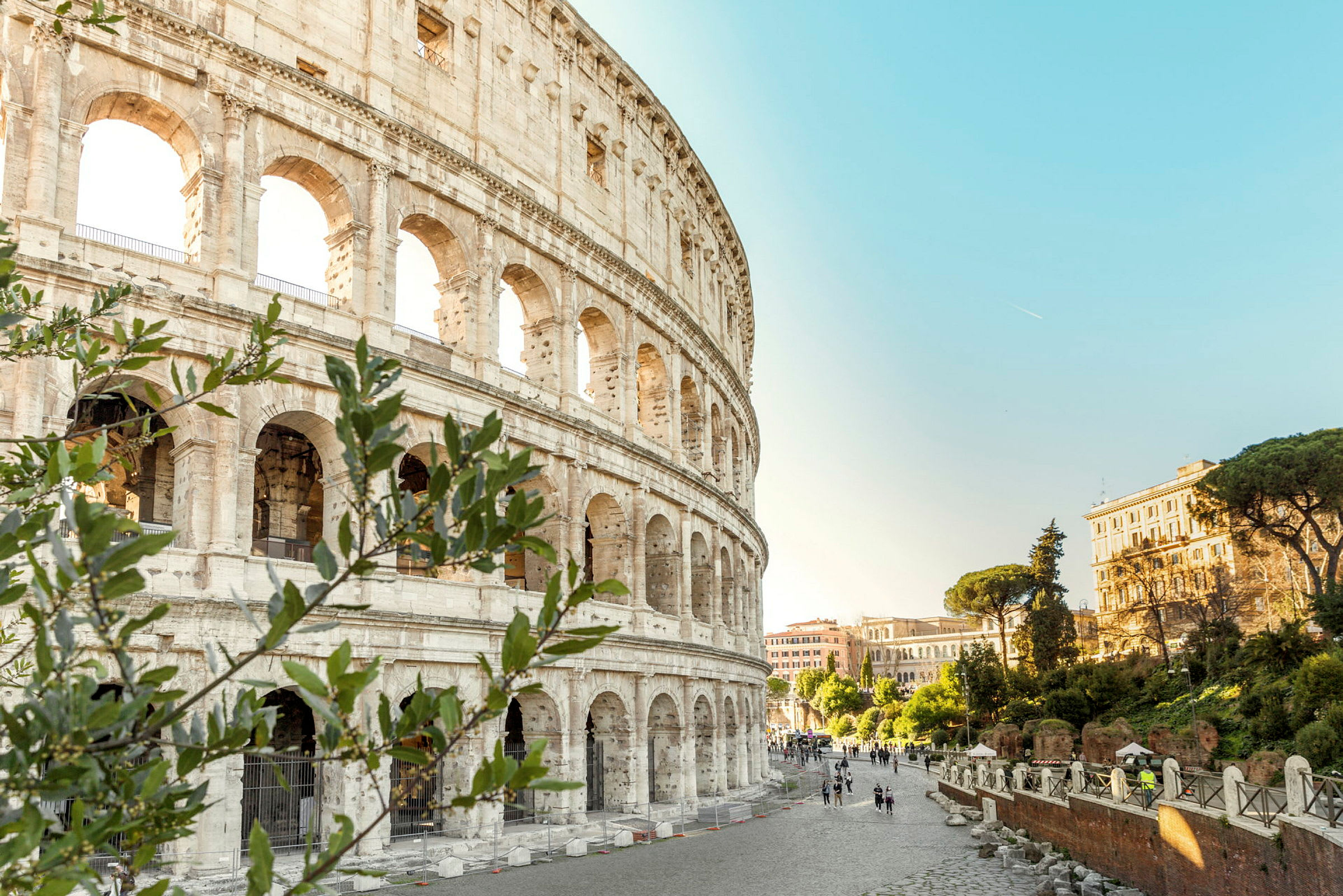 Billede af Colosseum med omliggende romersk arkitektur og grønne omgivelser under en klar blå himmel. (AI-genereret tekst)