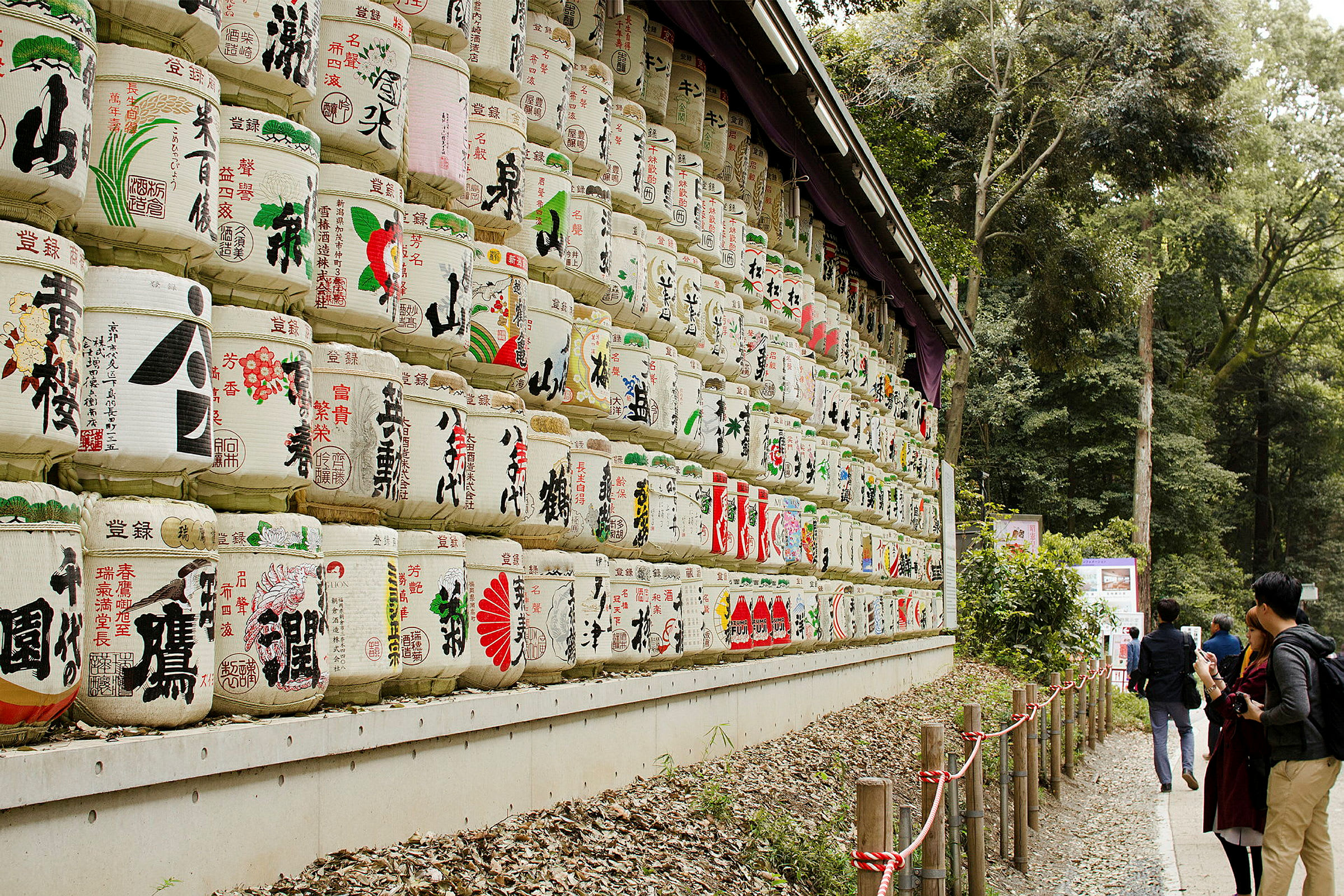 Et billede med stablede tønder dekoreret med japanske tegn og motiver, omgivet af en skov. Flere personer kigger på tønderne. (AI-genereret tekst)