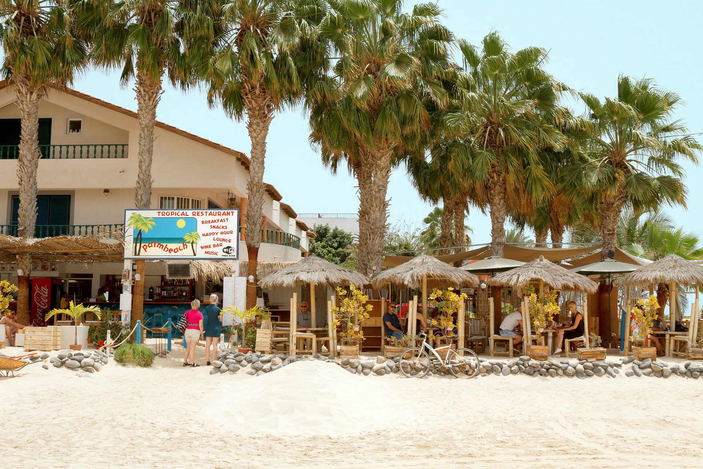 En tropisk restaurant med stråtag og palmer på en sandstrand. (AI-genereret tekst)