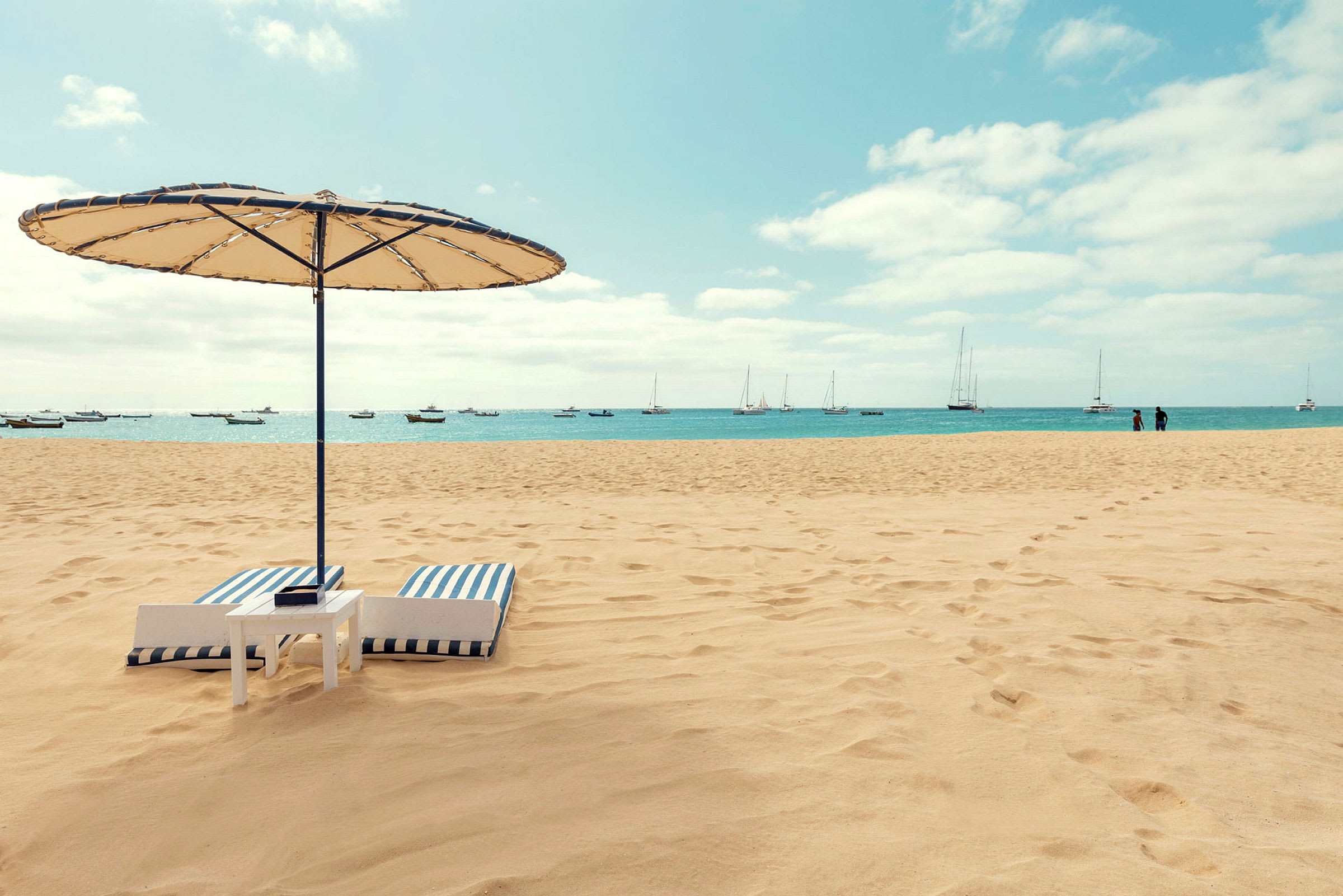 Billede af en strand med parasol, liggende solsenge og hav med både i baggrunden. (AI-genereret tekst)