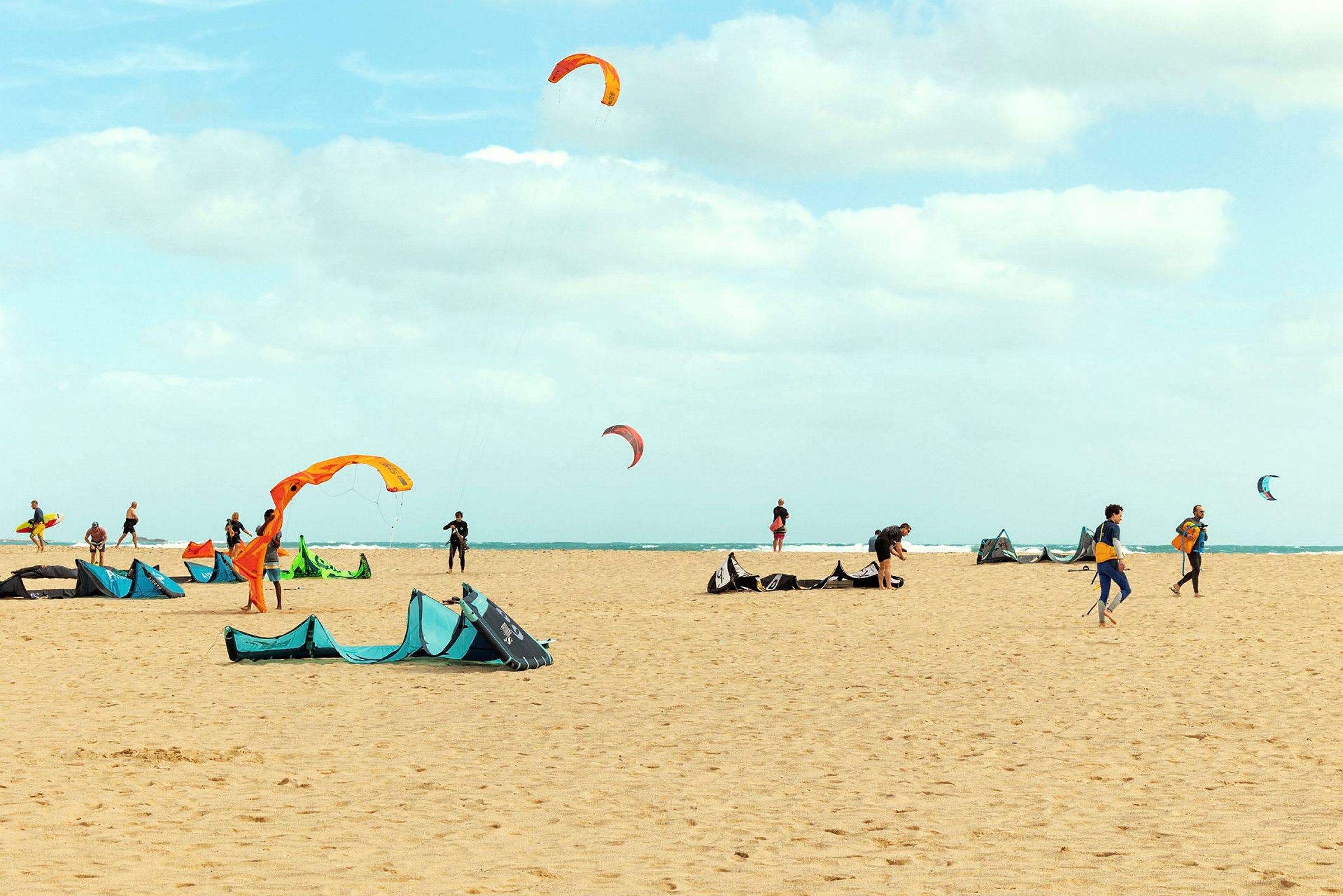 Strand med personer, der forbereder udstyr til kitesurfing med farverige drager i luften. (AI-genereret tekst)