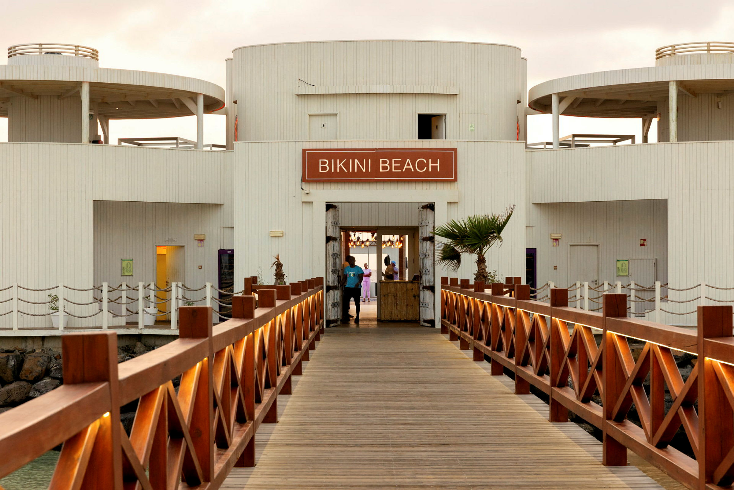 Udsigt over en træbro, der fører til en bygning med skiltet 'Bikini Beach'. (AI-genereret tekst)