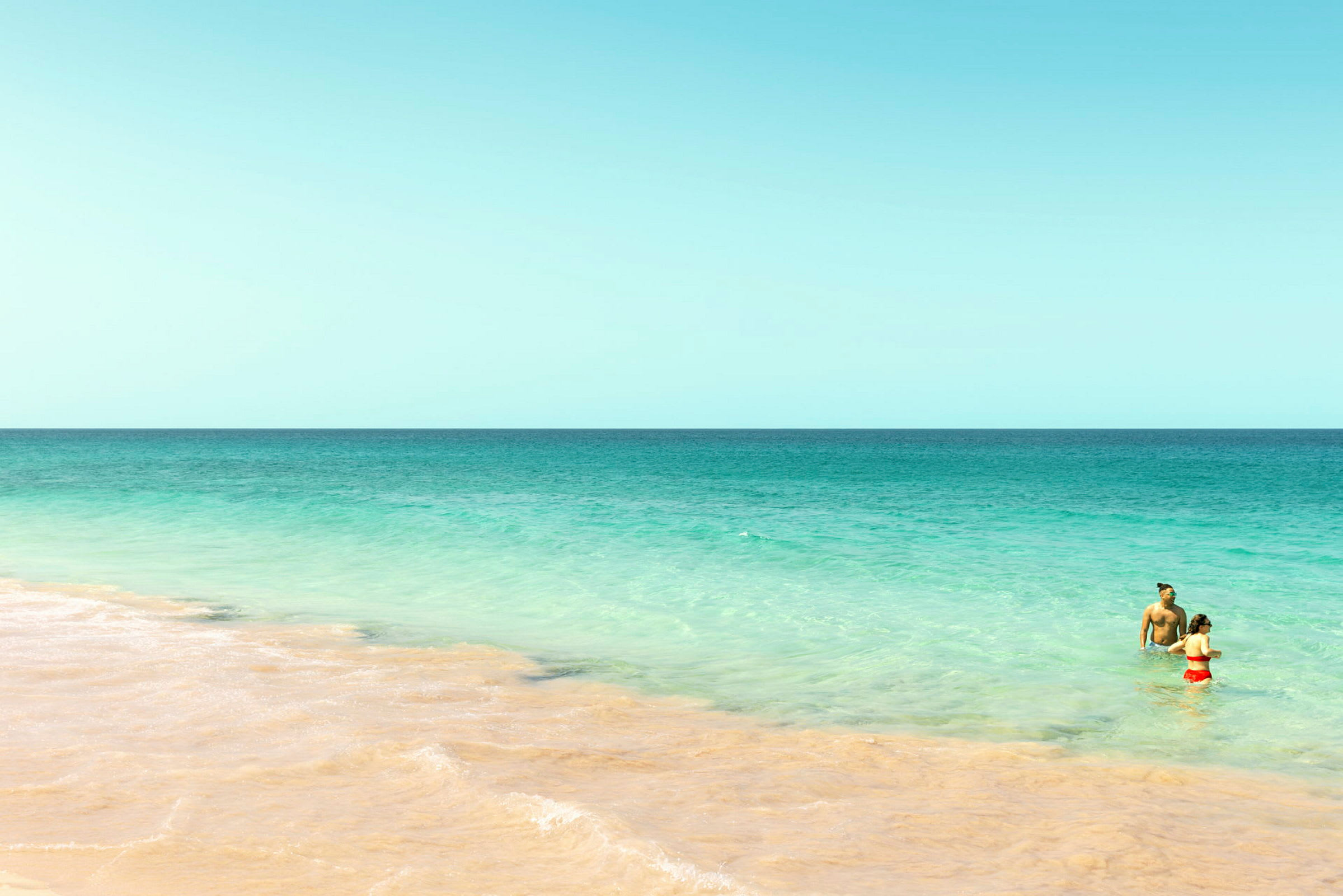 To personer står i det klare turkisblå hav nær en sandstrand under en skyfri himmel. (AI-genereret tekst)