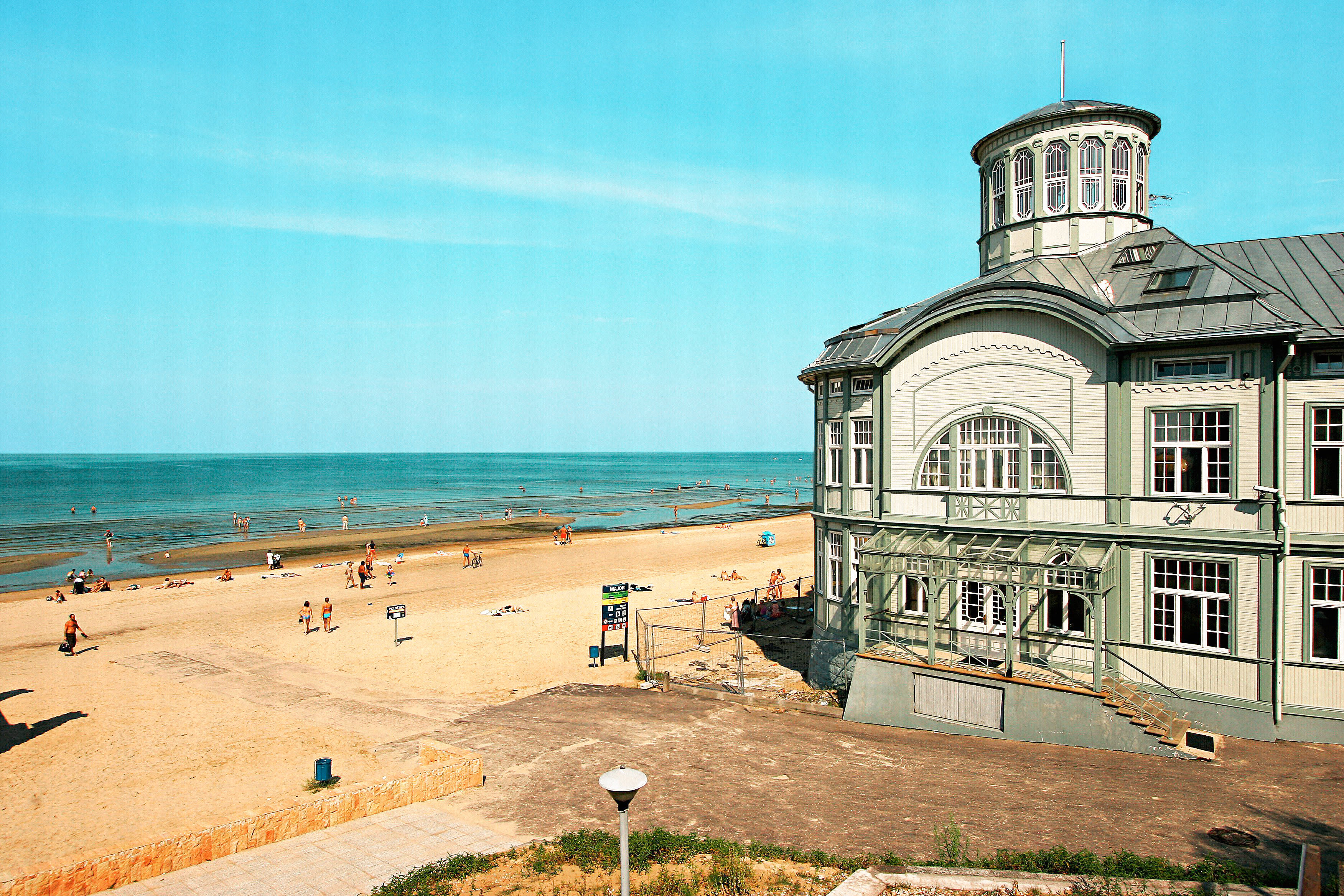 En strand med solbadende mennesker ved en rolig kystlinje, samt en større bygning med klassisk arkitektur ved stranden. (AI-genereret tekst)