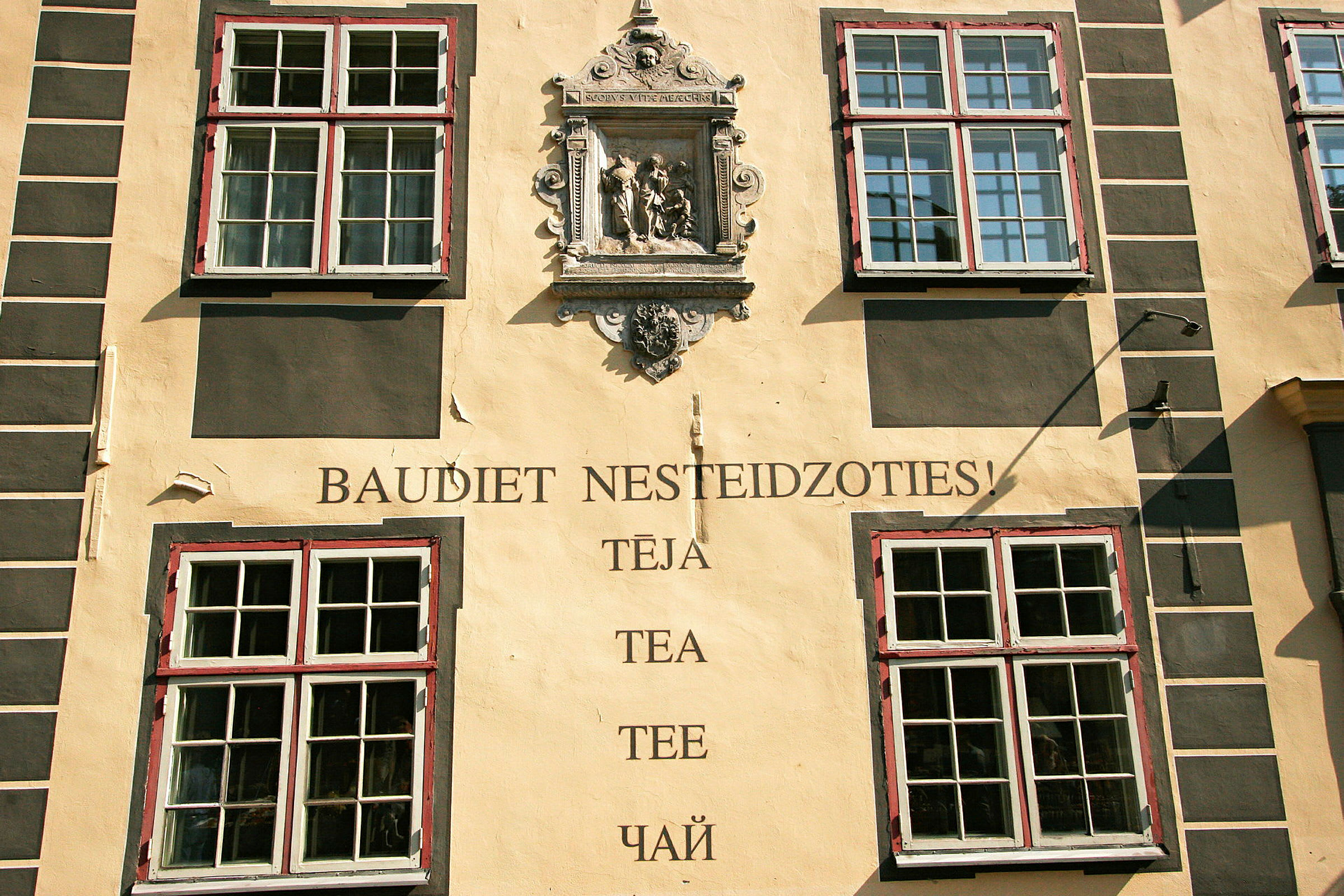 Facade af en bygning med fire store vinduer, en dekorativ stenrelief og teksten "Baudiet nesteidzoties! Tēja, Tea, Tee, Чай". (AI-genereret tekst)
