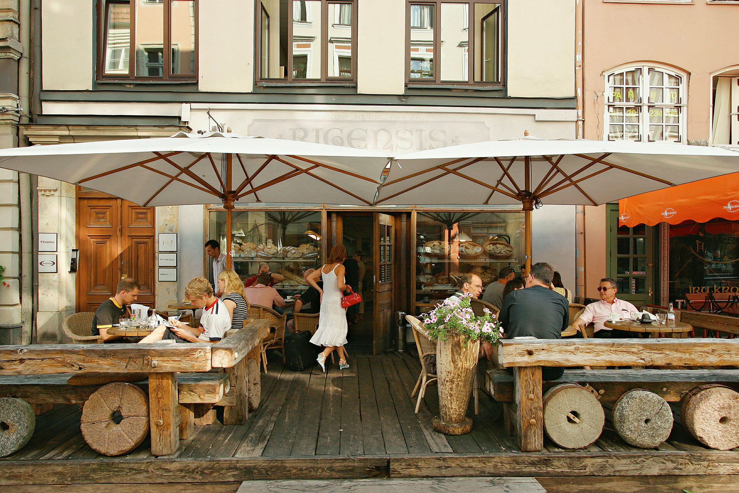 Et café med gæster, der sidder udendørs ved rustikke træborde under store parasoller. (AI-genereret tekst)