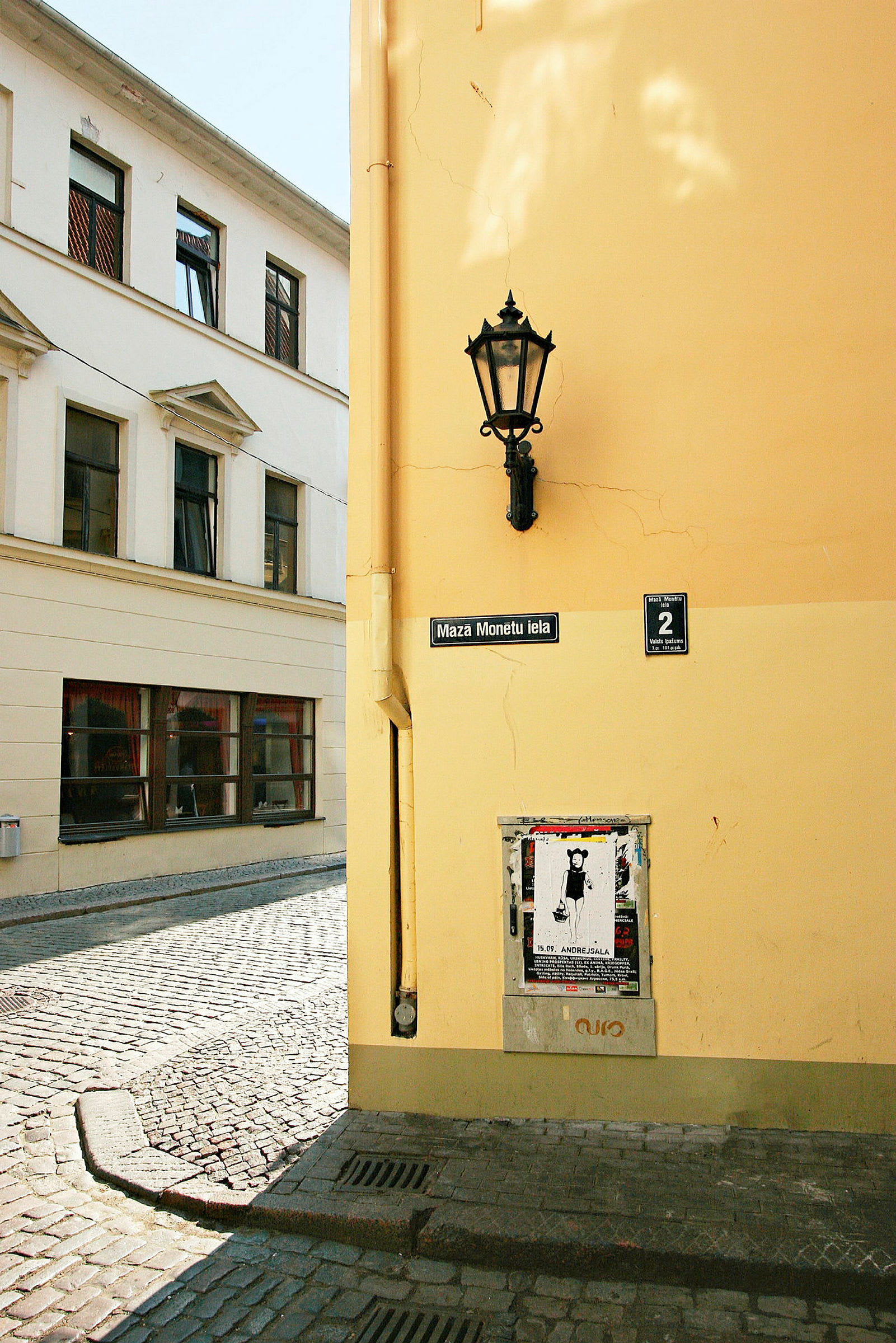 Et hjørne af en bygning med gul facade, en gadelampe, et skilt med teksten 'Maza Monētu iela' og et område med plakater. (AI-genereret tekst)