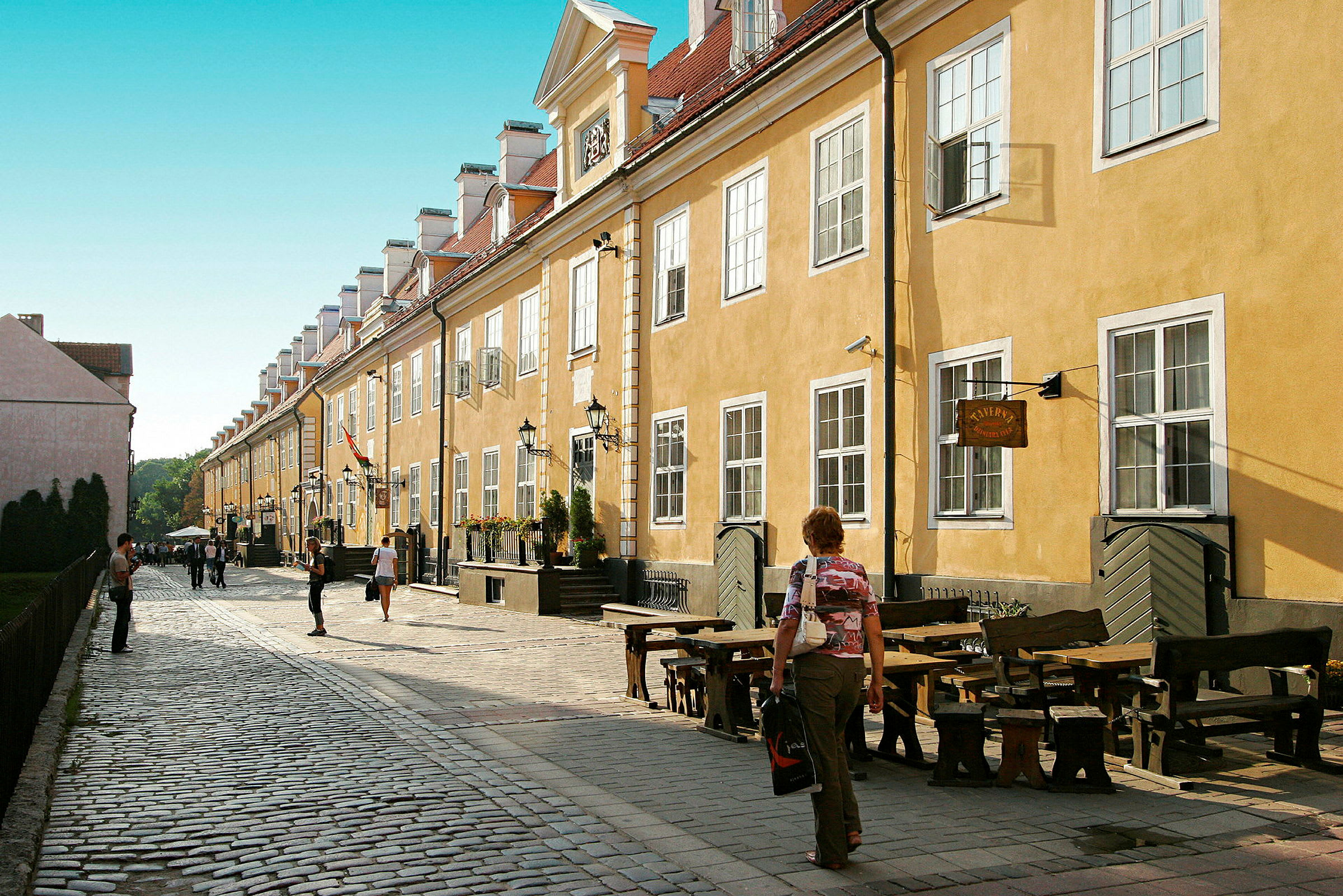 En brostensbelagt gade omgivet af gule bygninger med hvide vinduer og nogle udendørs serveringsområder. (AI-genereret tekst)
