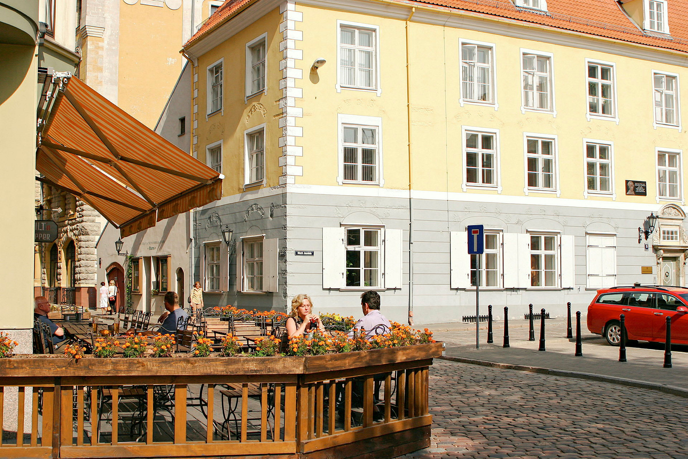 Gadebillede med udendørs servering omgivet af blomster og historiske bygninger. (AI-genereret tekst)